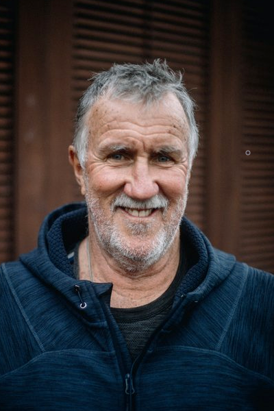 A smiling middle-aged man with gray hair and a beard, wearing a navy blue hoodie, standing outdoors in front of a wooden background.