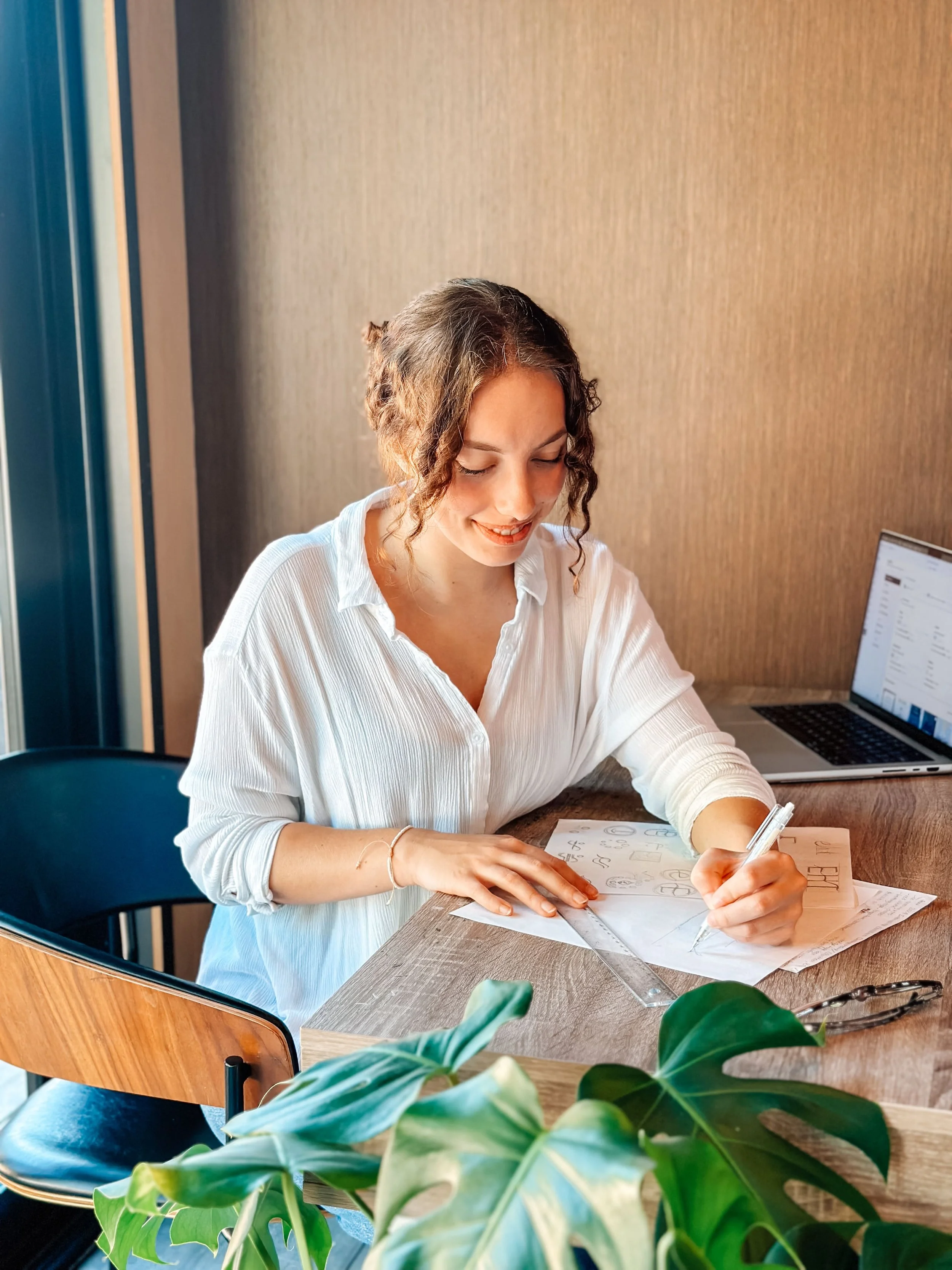 A woman sitting at a desk, working on sketches with a pen, papers, a ruler, a laptop, and a plant nearby.