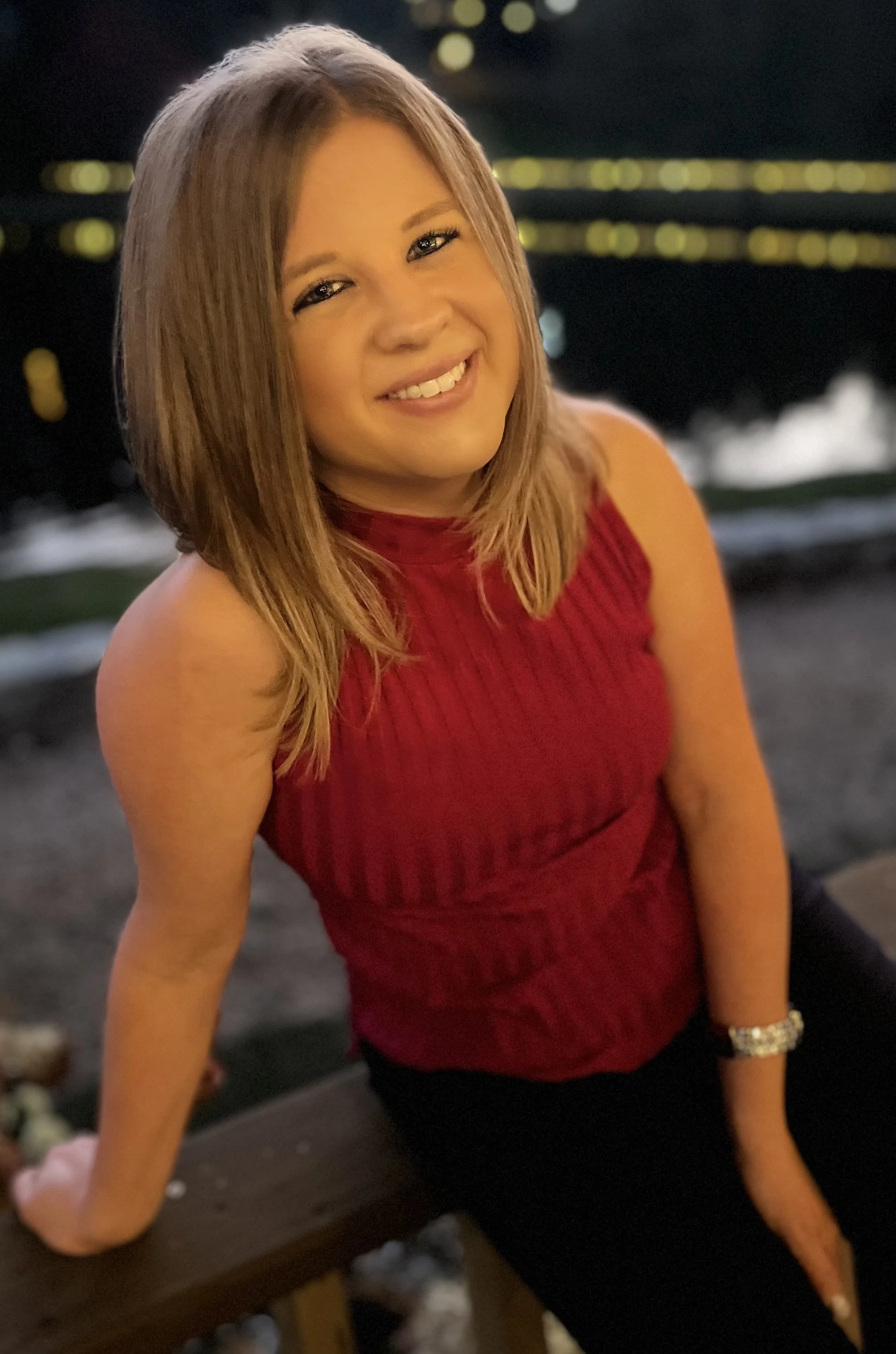 Taylor Capps, a woman with shoulder-length light brown hair smiling, wearing a sleeveless red top and sitting on a wooden surface at night with blurred city lights in the background.