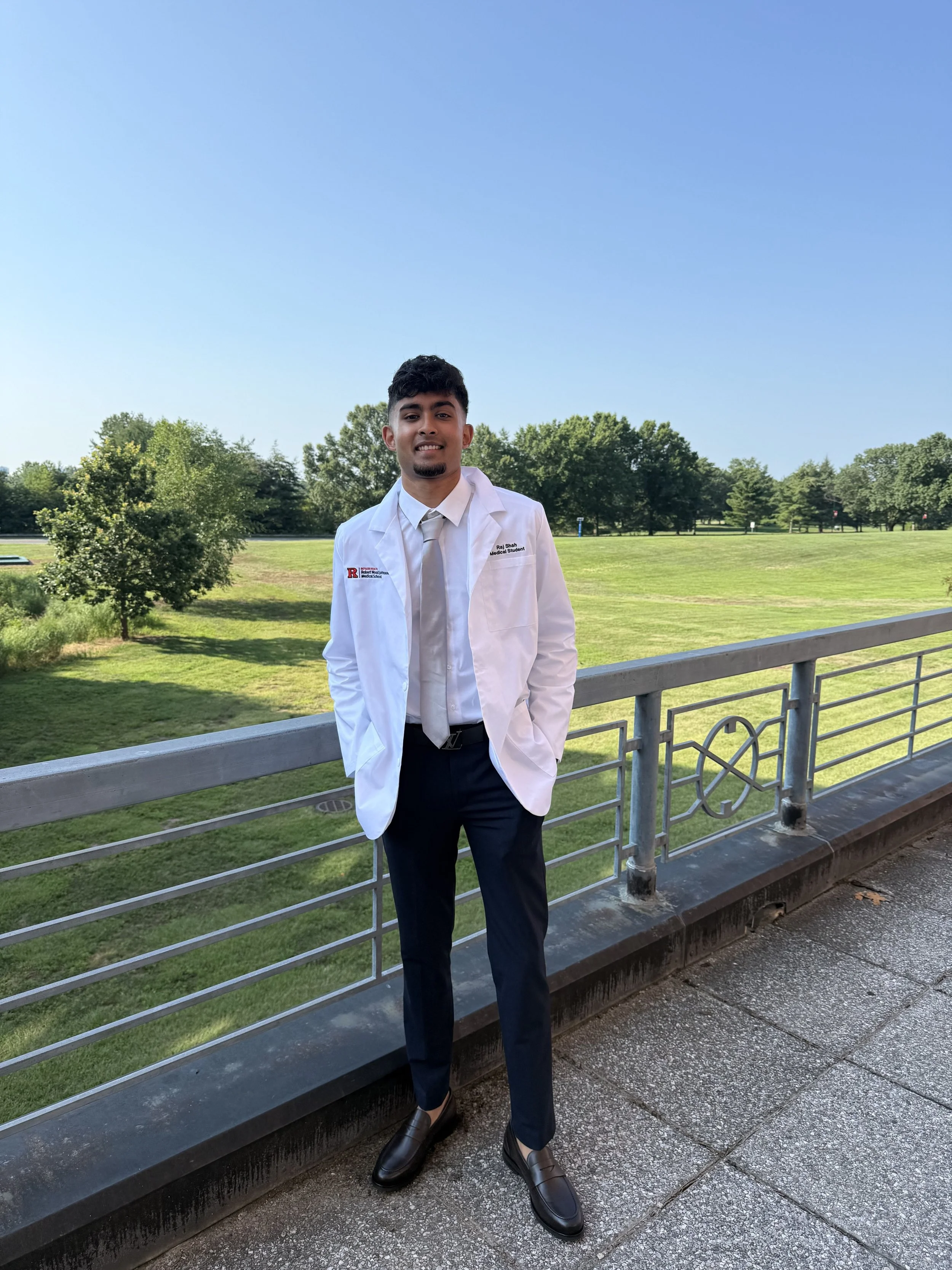 Young man in a white medical coat standing on a balcony with a park in the background during daytime.