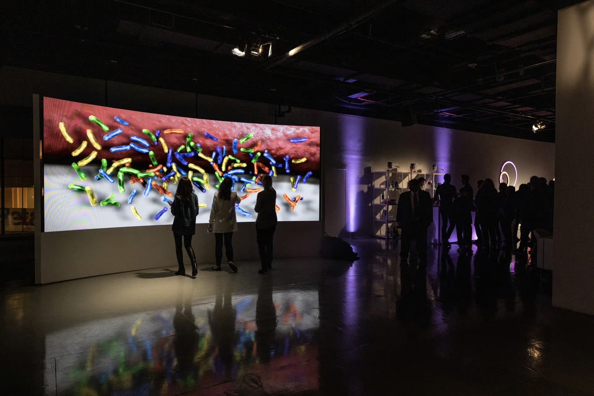 An indoor exhibition with a large digital screen displaying colorful microscopic images; people are gathered in front of the screen, observing and interacting with the display.