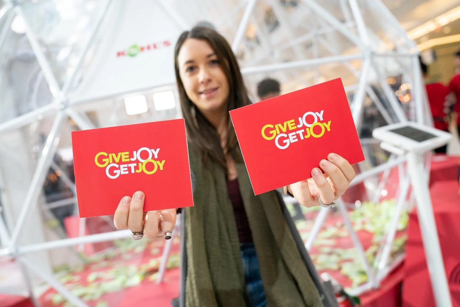 Woman holding red cards with yellow and white text that reads "GIVE JOY GET JOY" at an indoor event or market.