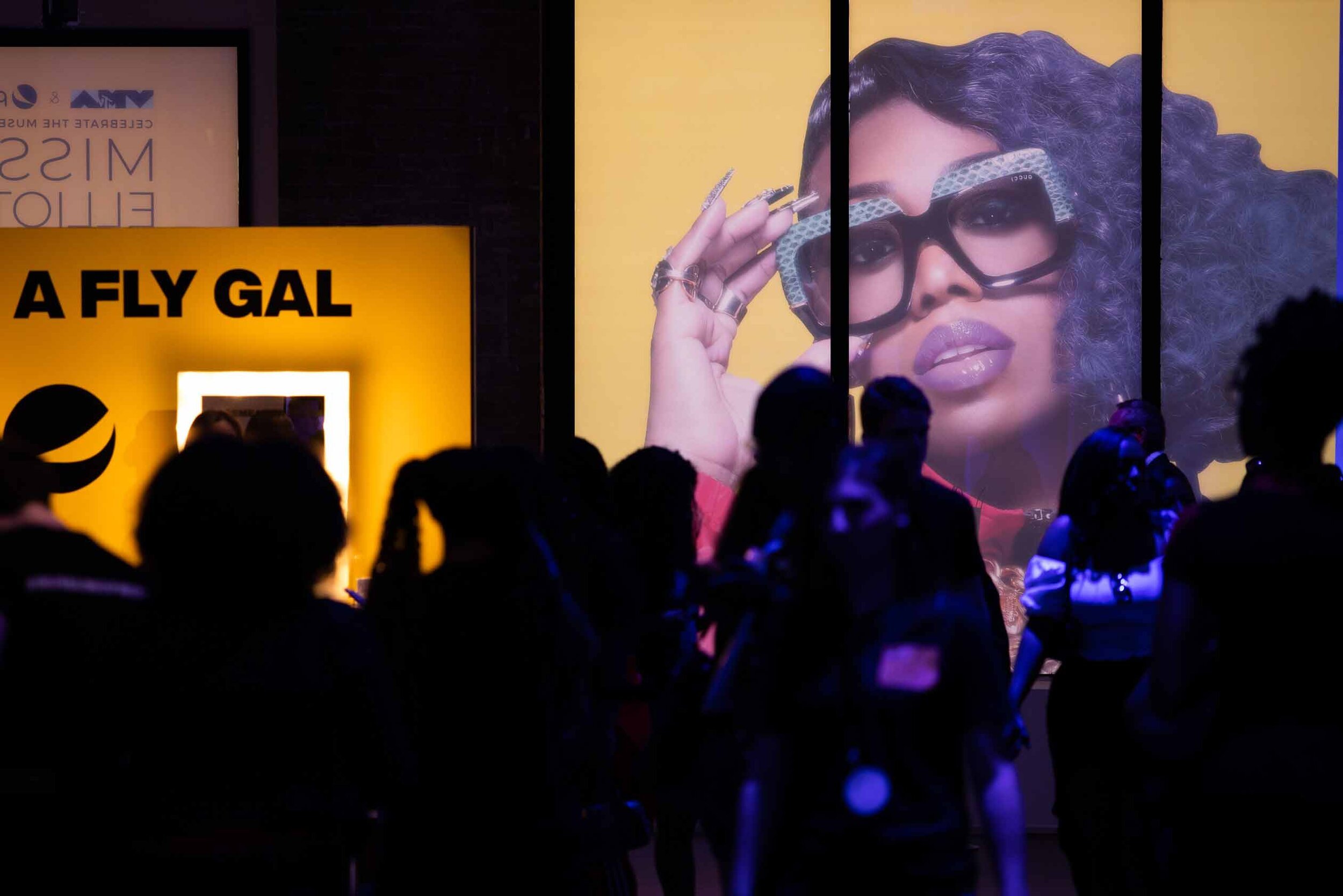 People standing in front of a large billboard featuring a woman with curly hair, wearing glasses, and holding her hand near her forehead. The billboard has a yellow background. To the left, there is a yellow structure with the text 'A FLY GAL' and a 