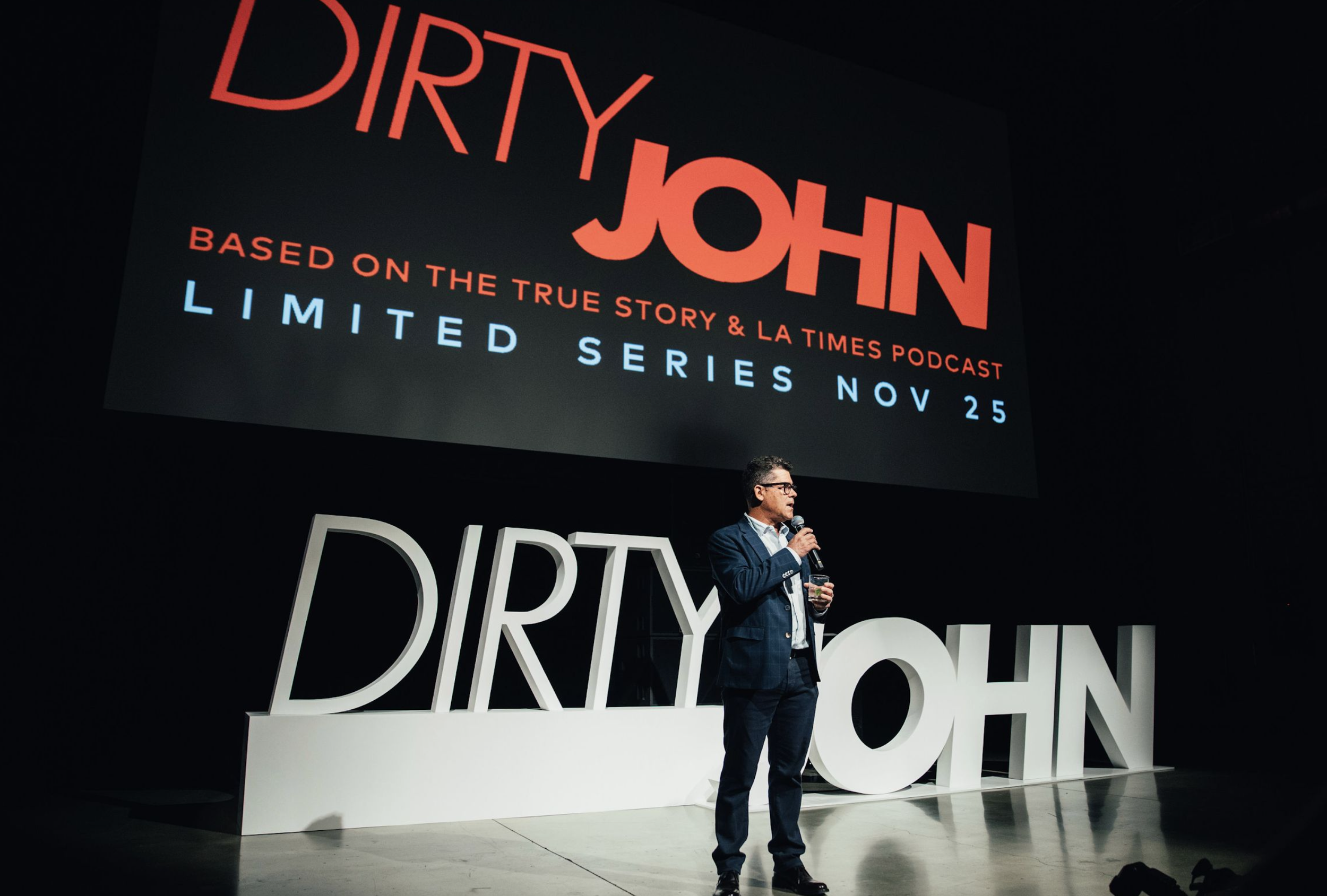 A man in a suit holding a microphone presents on stage in front of large 3D letters spelling 'DIRTY JOHN' and a screen displaying promotional text for a series based on a podcast, with release date November 25.