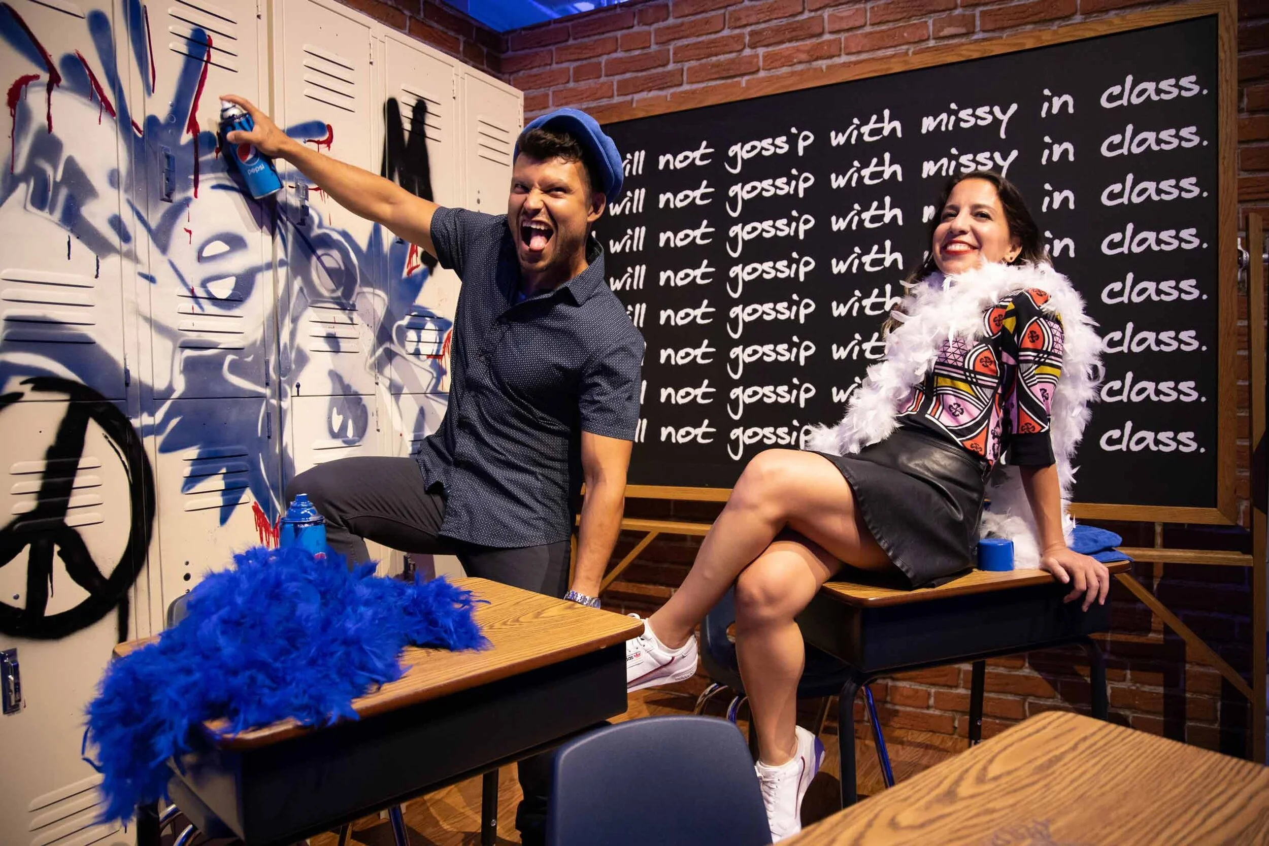 Two people in a classroom decorated with graffiti and lockers, one man spraying graffiti on lockers and the woman sitting on a desk, smiling; a blackboard in the background with the phrase 'I will not gossip with missy in class' written multiple time