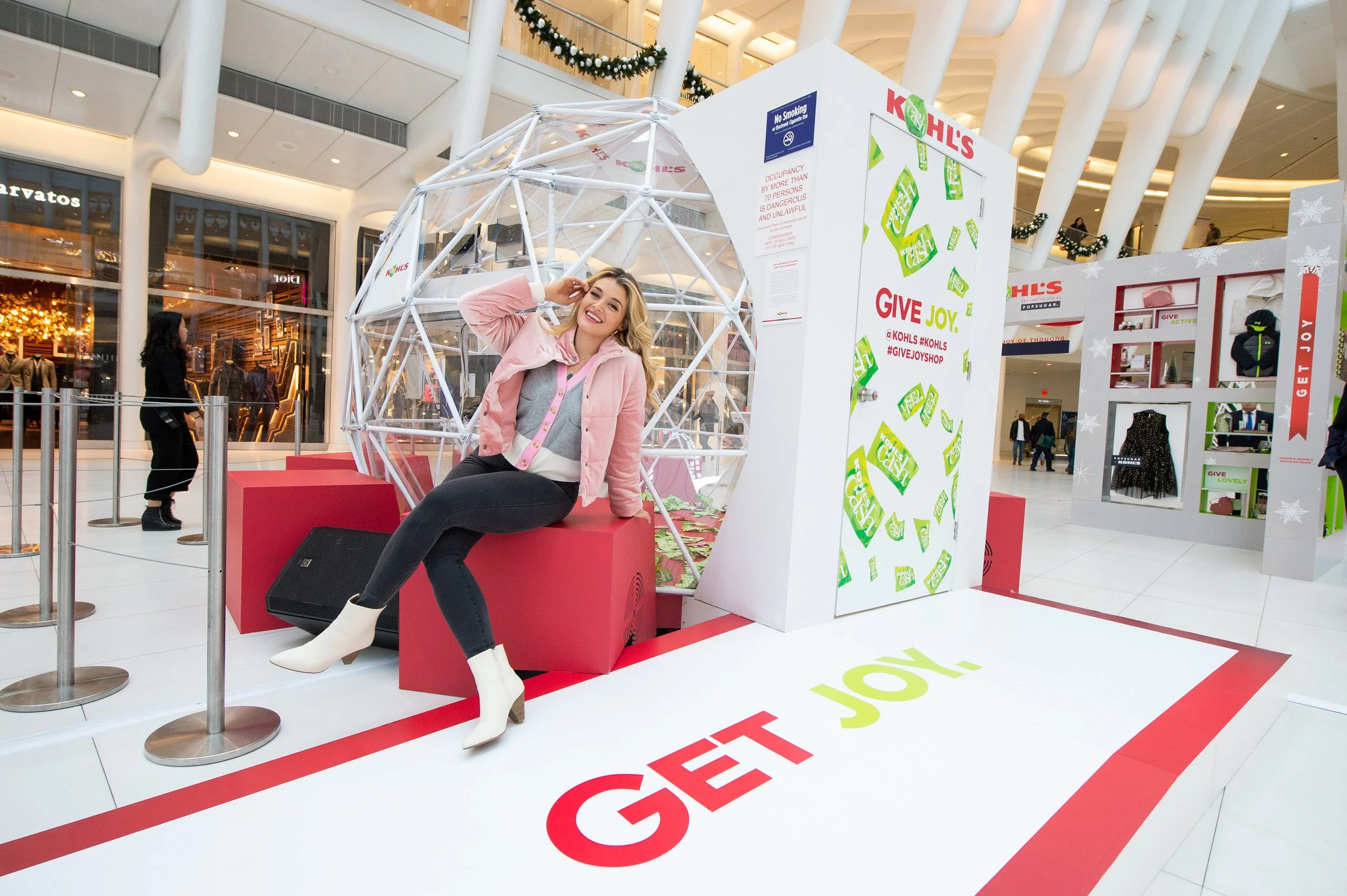 A young woman sitting on a red platform inside a shopping mall, smiling and posing with one hand touching her head, in front of a large white cube display with green cash-like graphics and the words "GIVE JOY." The scene is decorated for Christmas wi