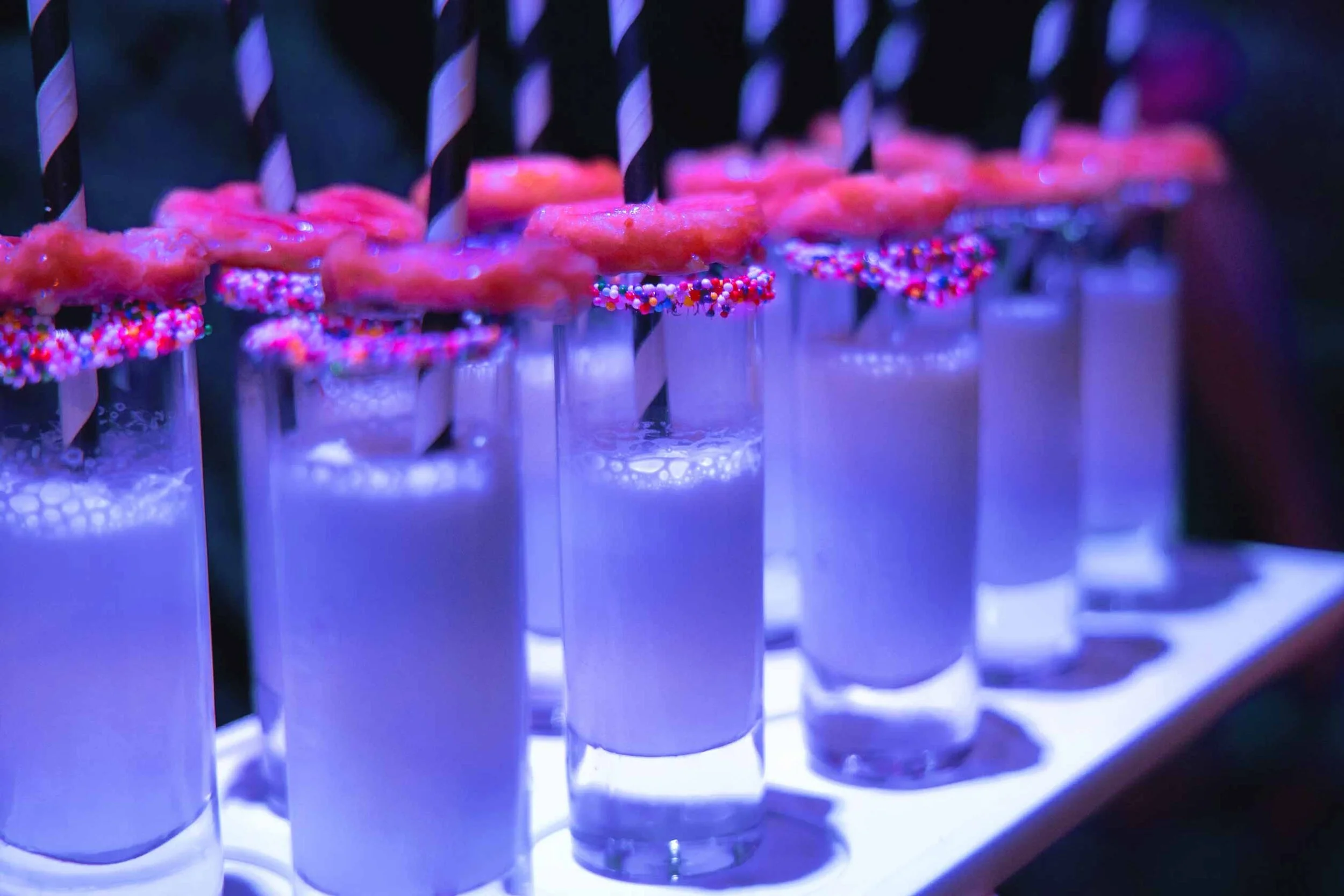 Row of milkshakes in tall glasses with frosting and sprinkles, topped with donuts on striped straws under blue lighting.