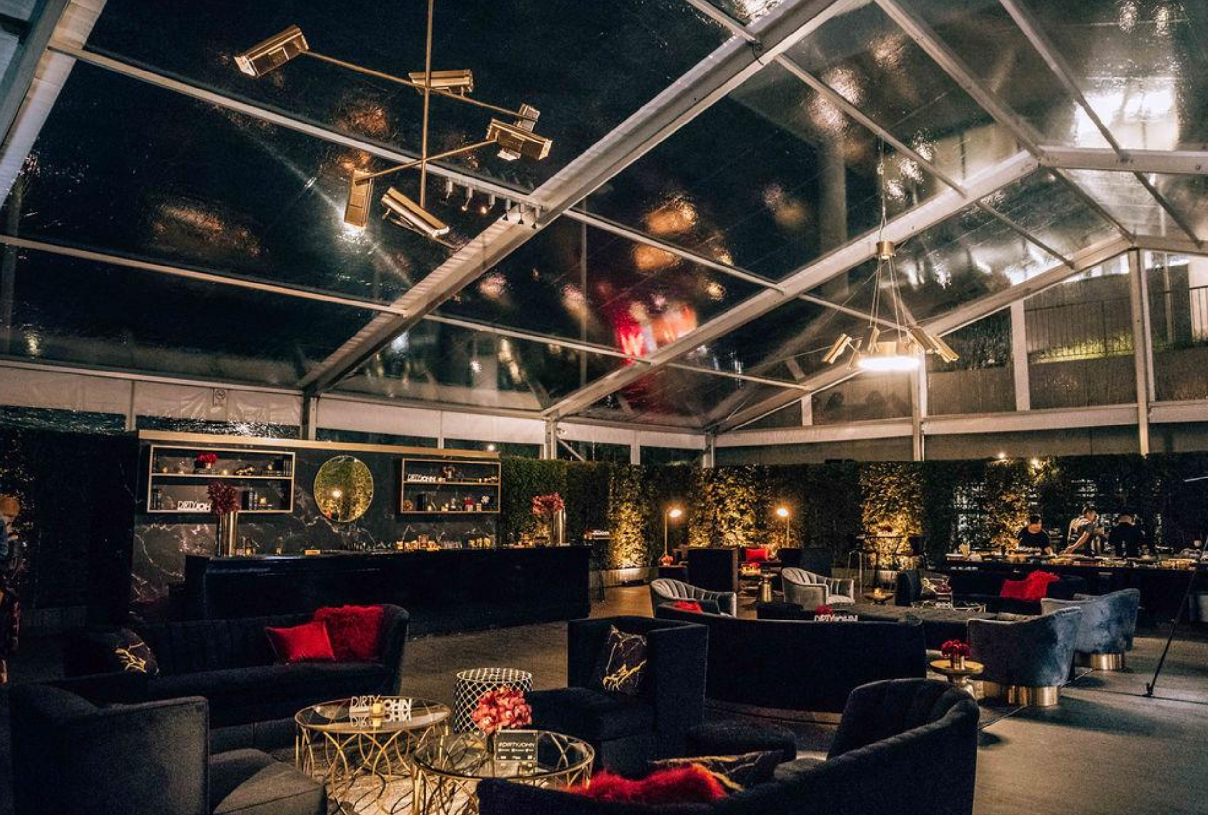 Interior of a modern, upscale lounge or event space with black velvet couches, gold accents, a high glass ceiling, and warm lighting at night.