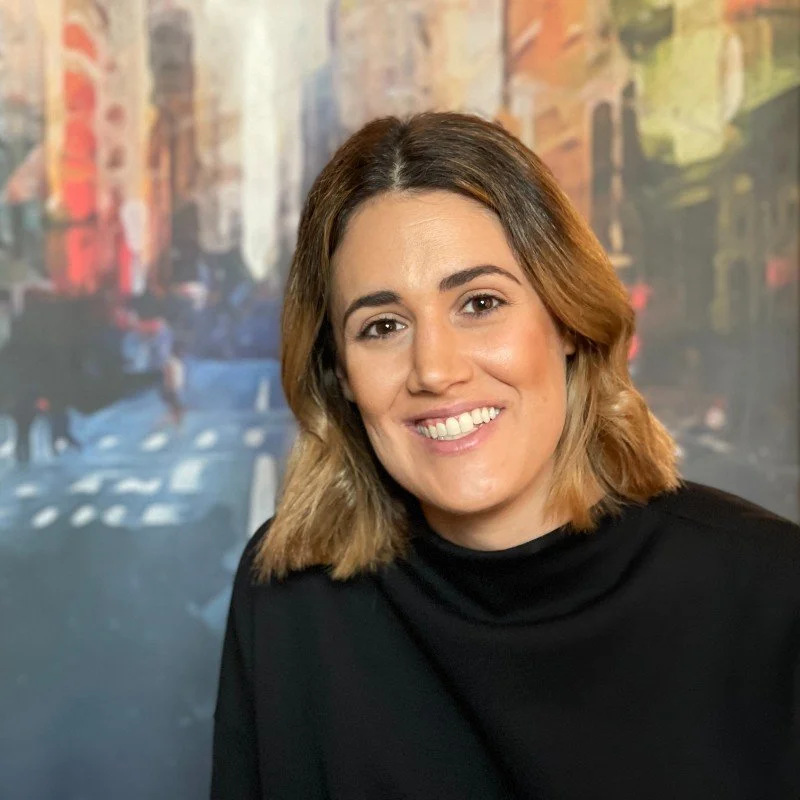 A woman with shoulder-length brown hair, smiling, wearing a black top, with a colorful cityscape painting or mural in the background.