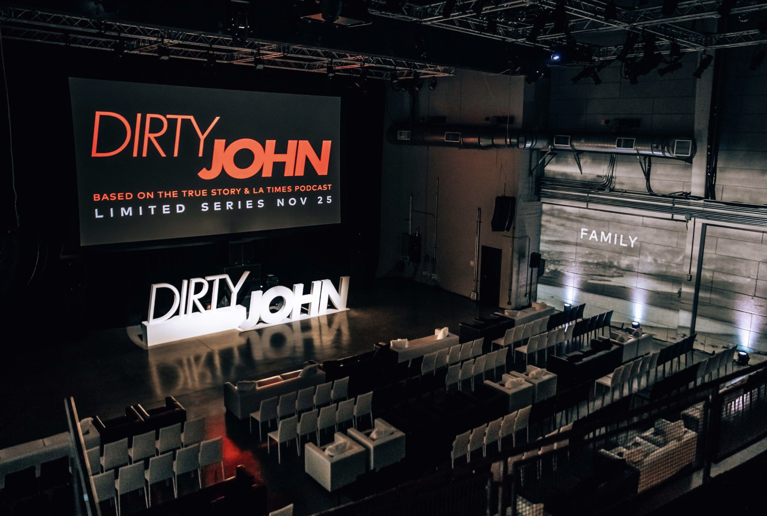 An indoor event space set up for a presentation or performance, featuring a large screen displaying advertisements for the 'Dirty John' podcast and limited series, with seating arranged in rows and a prominent illuminated sign that reads 'DIRTY JOHN'