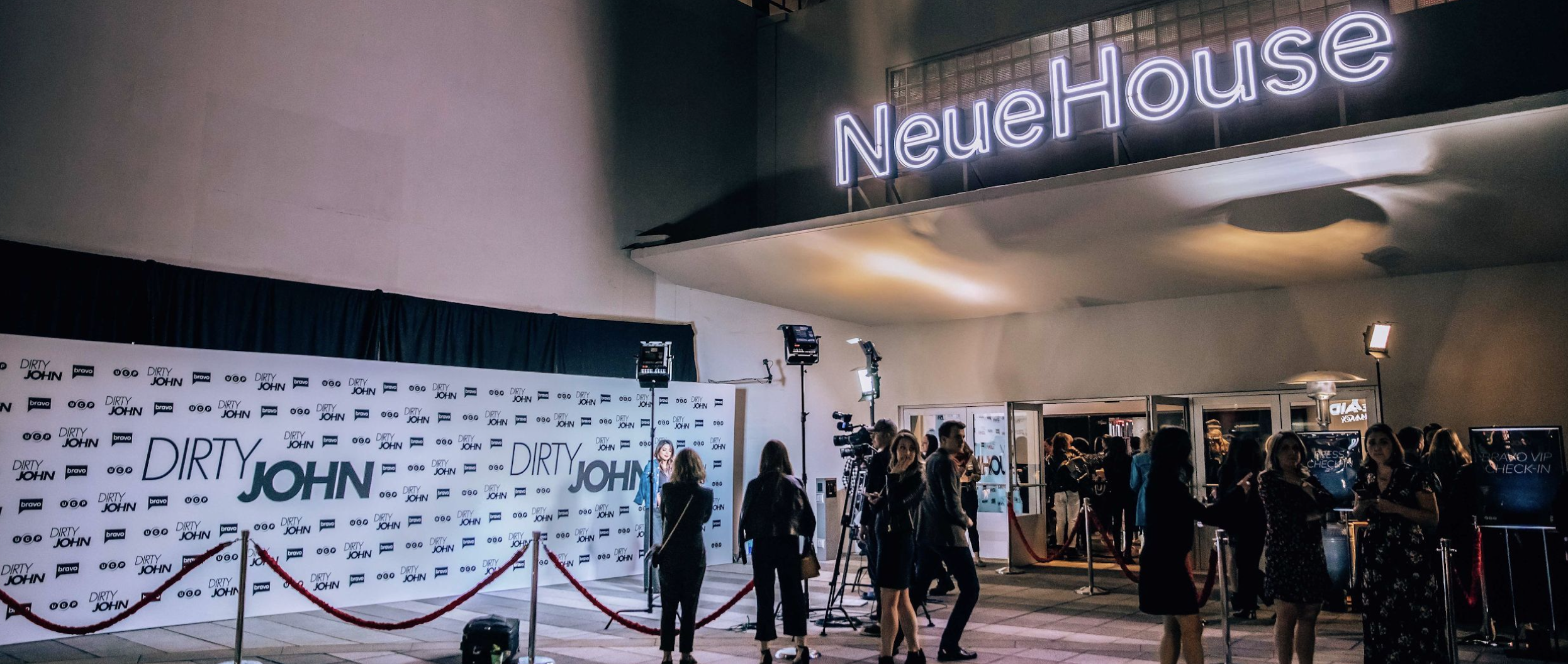 People waiting in line outside of a venue called NeueHouse, with a backdrop that says 'Dirty John' and cameras set up for filming, under a neon sign.