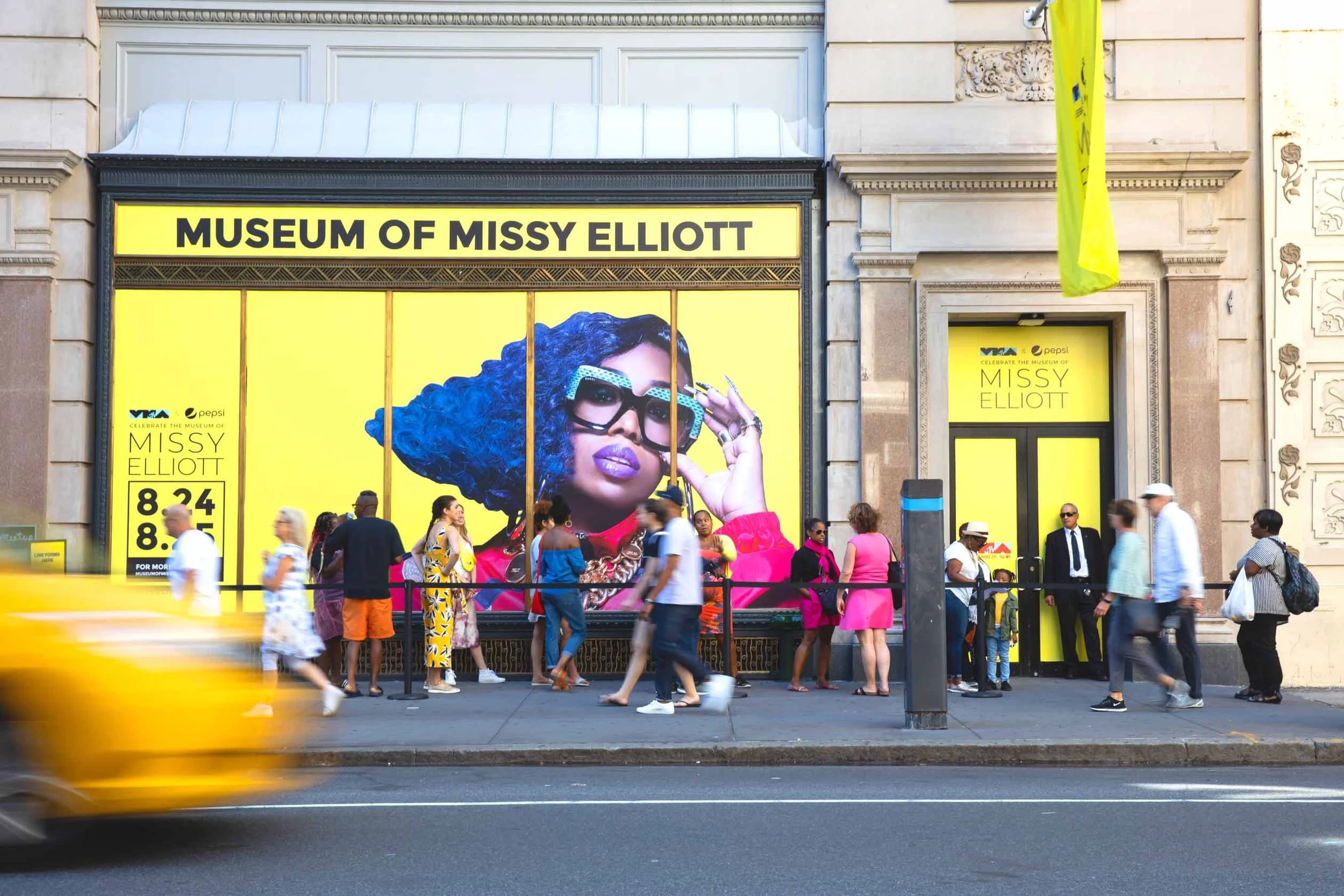 Exterior of the Museum of Missy Elliott with large mural of Missy Elliott in colorful outfit and glasses, people walking in front, yellow signage, and storefront entrance.