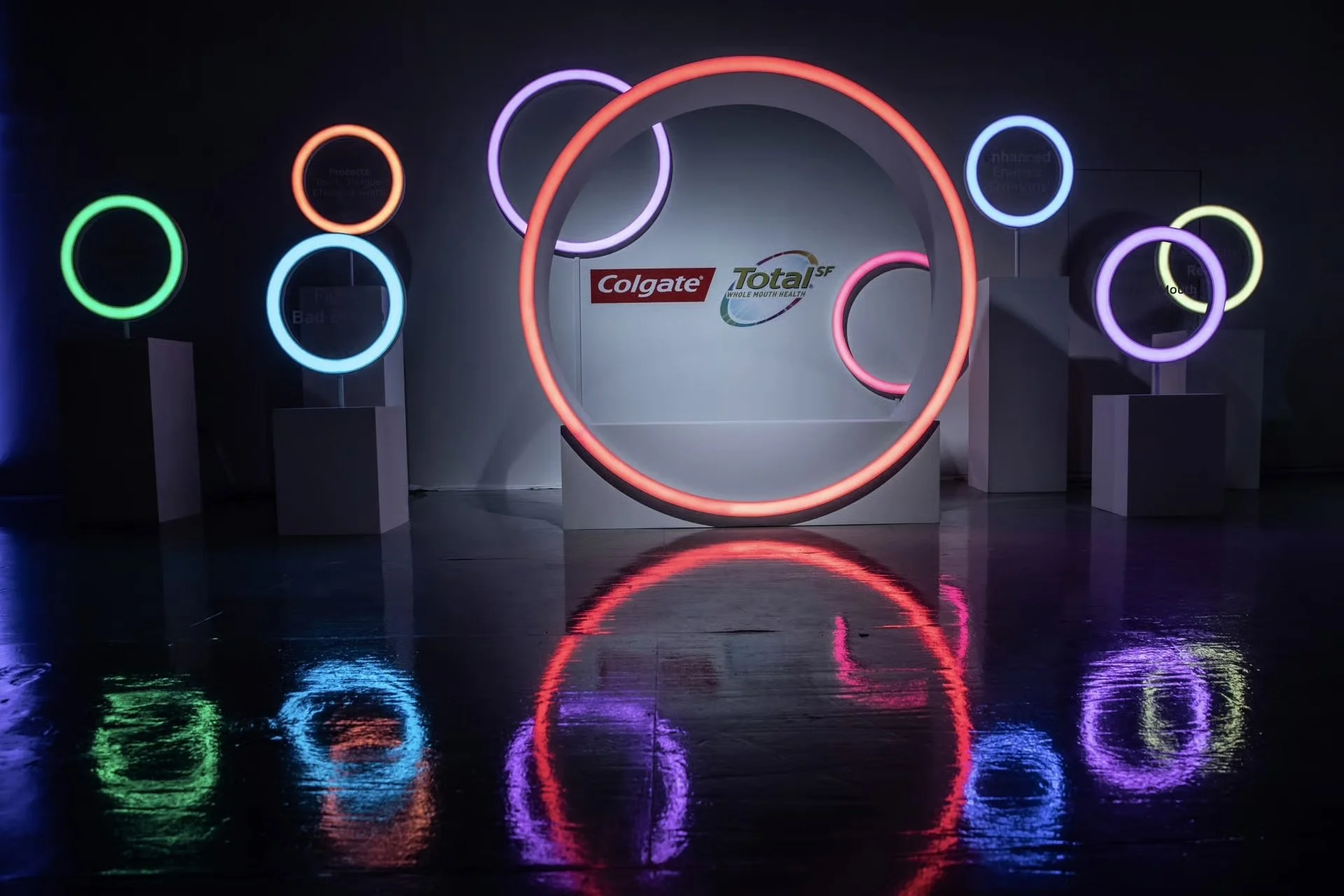 Colorful neon rings of various sizes illuminated in a dark room, with their reflections visible on the shiny floor, and a white backdrop with Colgate and Total logos in the background.
