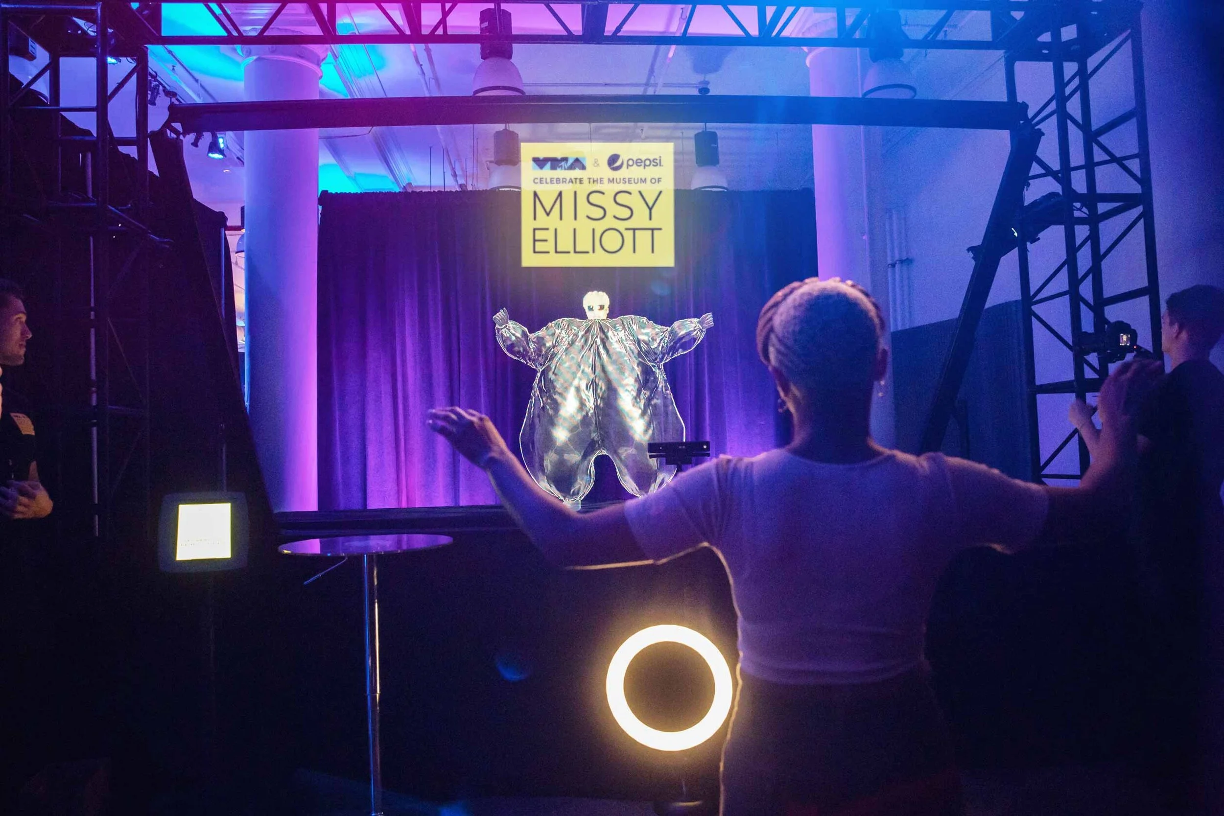 A person with arms outstretched facing a stage with a purple curtain, a figure dressed in a shiny outfit, and a sign that reads 'Celebrate the Museum of Missy Elliott' at an event or exhibit.