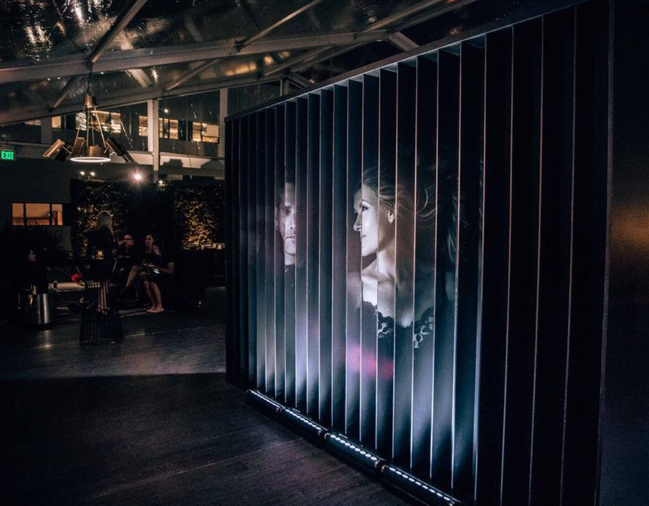 Interior of a modern art gallery or an event space with a large illuminated portrait of two women on a partition. The space is dimly lit with a few people sitting at tables in the background.