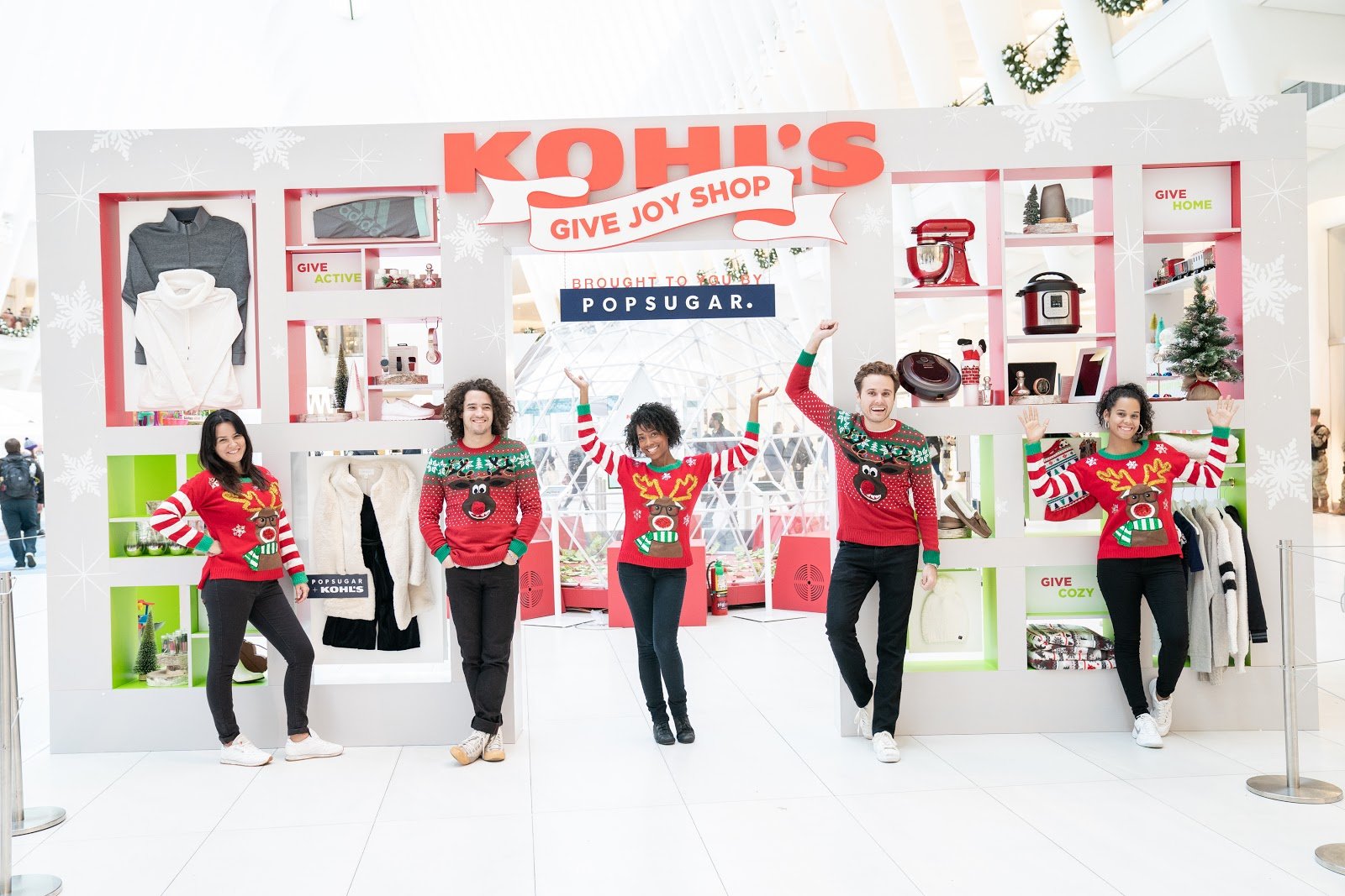 People wearing Christmas sweaters standing in front of a holiday-themed Kohl's pop-up shop display inside a shopping mall.