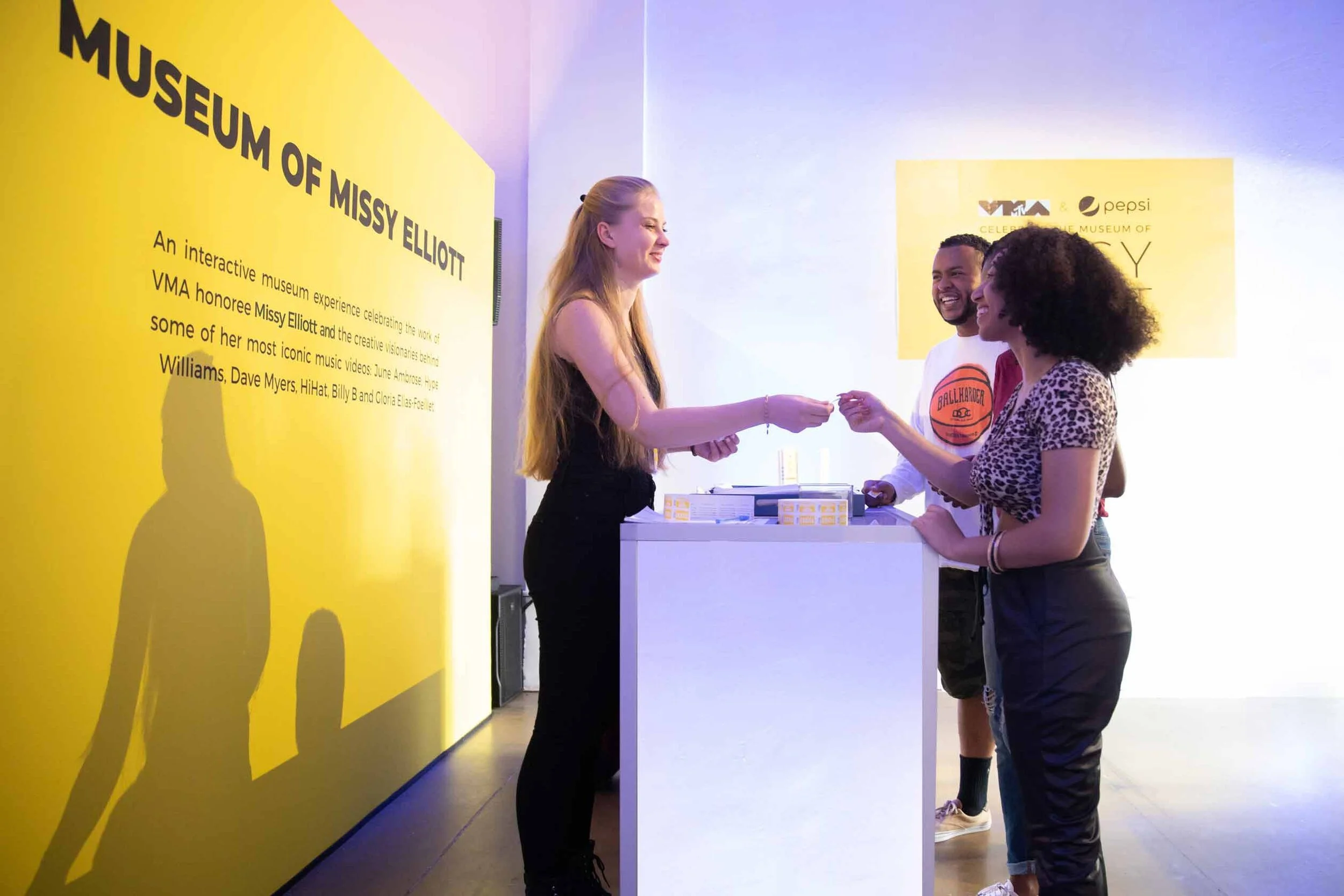 A woman with long blonde hair handing something to a woman with curly hair at an event with yellow walls and informational signs.