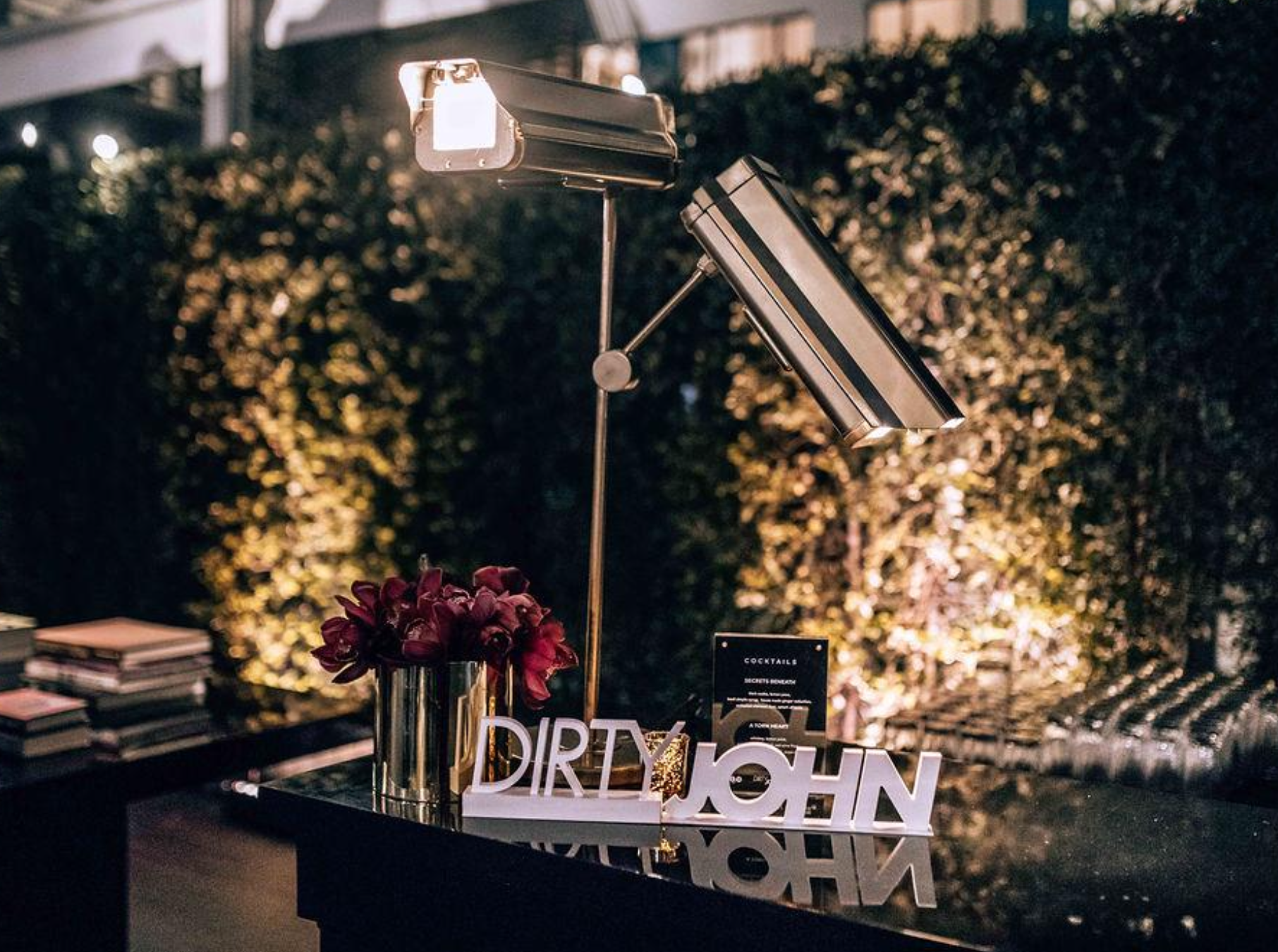A modern outdoor bar setup with a bouquet of dark pink flowers, illuminated signage spelling 'DIRTY JOHN', a black menu card, and stylized metal lamps, surrounded by greenery at night.