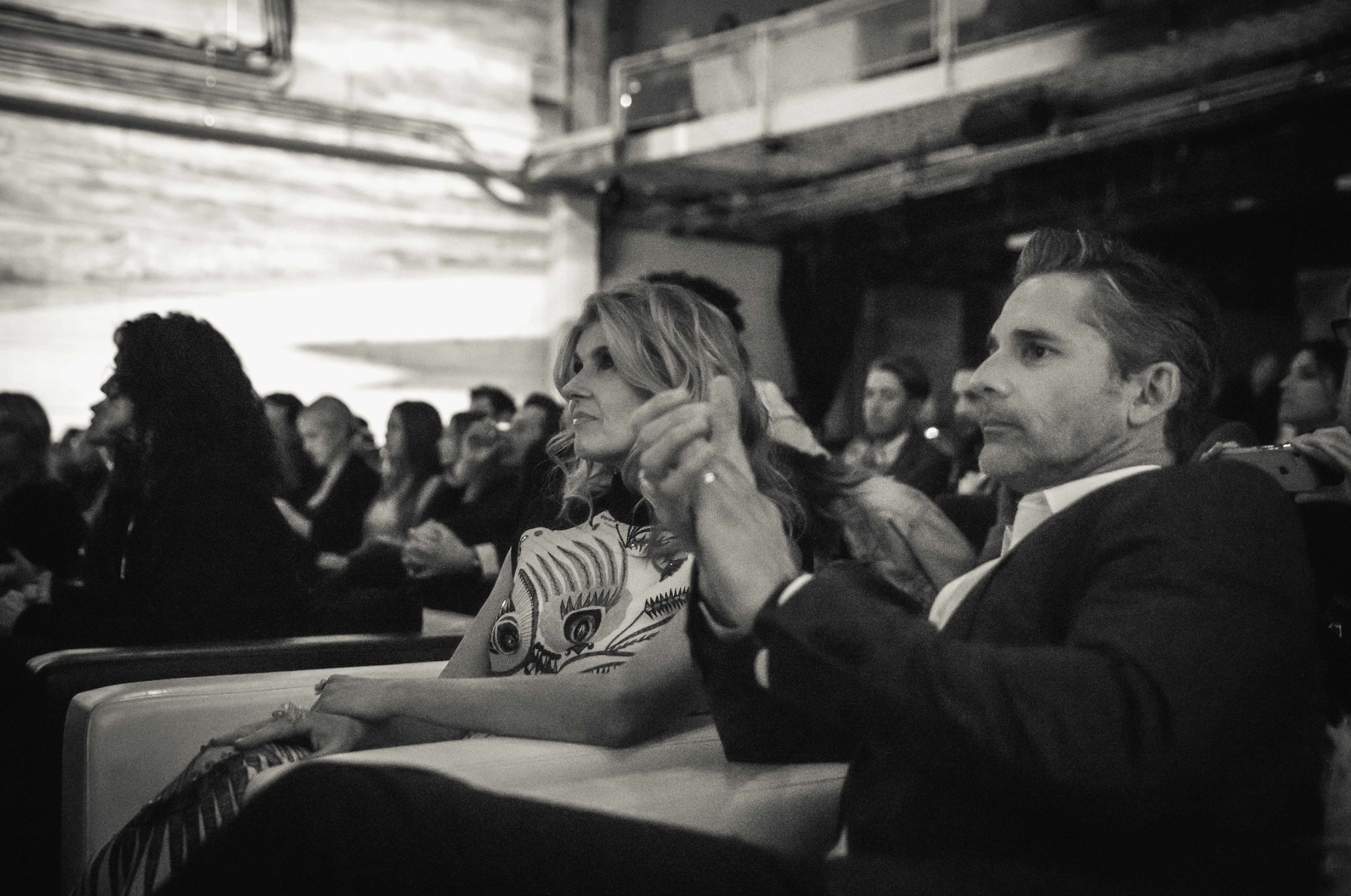 A black and white photo of an audience sitting in a theater or conference hall, focusing on a woman with blonde wavy hair and a man with dark, slicked-back hair, both attentively watching the stage.