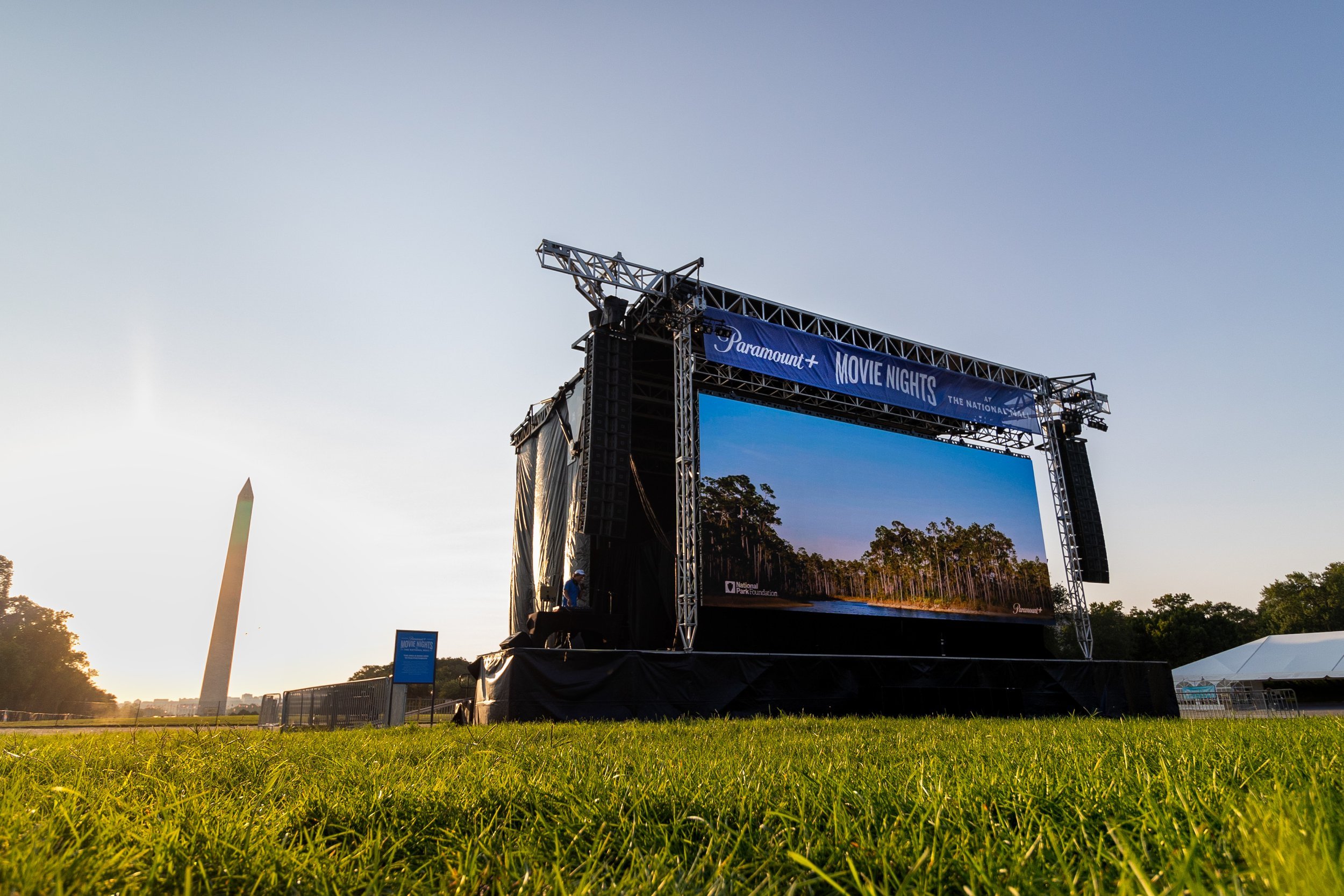 Outdoor stage with large screen and banner reading 'Paramount+ Movie Nights.' The stage is set on a grassy field with trees and a monument in the distance, under a clear sky.