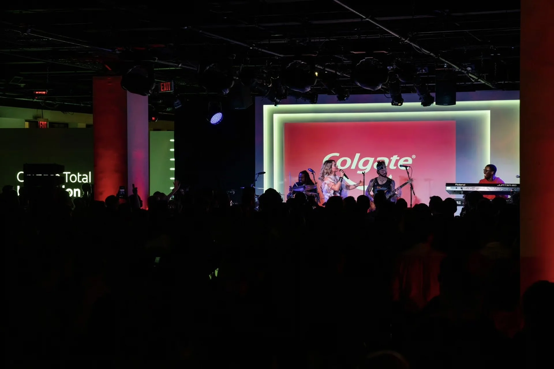 A live music performance on stage at an event sponsored by Colgate, with a female singer, a keyboardist, and two musicians playing drums and guitar, in front of an audience.