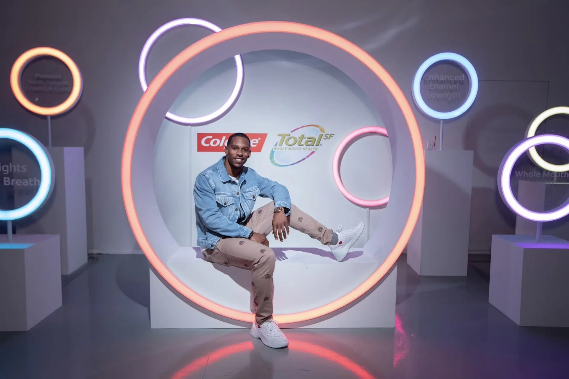 A man sitting inside a neon-lit circular art display at an exhibit, smiling, wearing a denim jacket, beige pants, and white sneakers, with logos and colorful neon rings in the background.