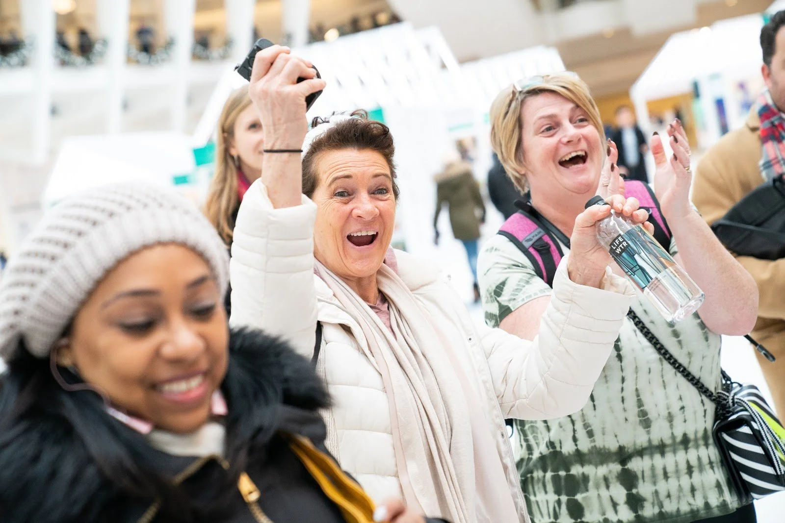 Group of people at an indoor event, smiling and cheering, with one woman holding a camera and another drinking water.