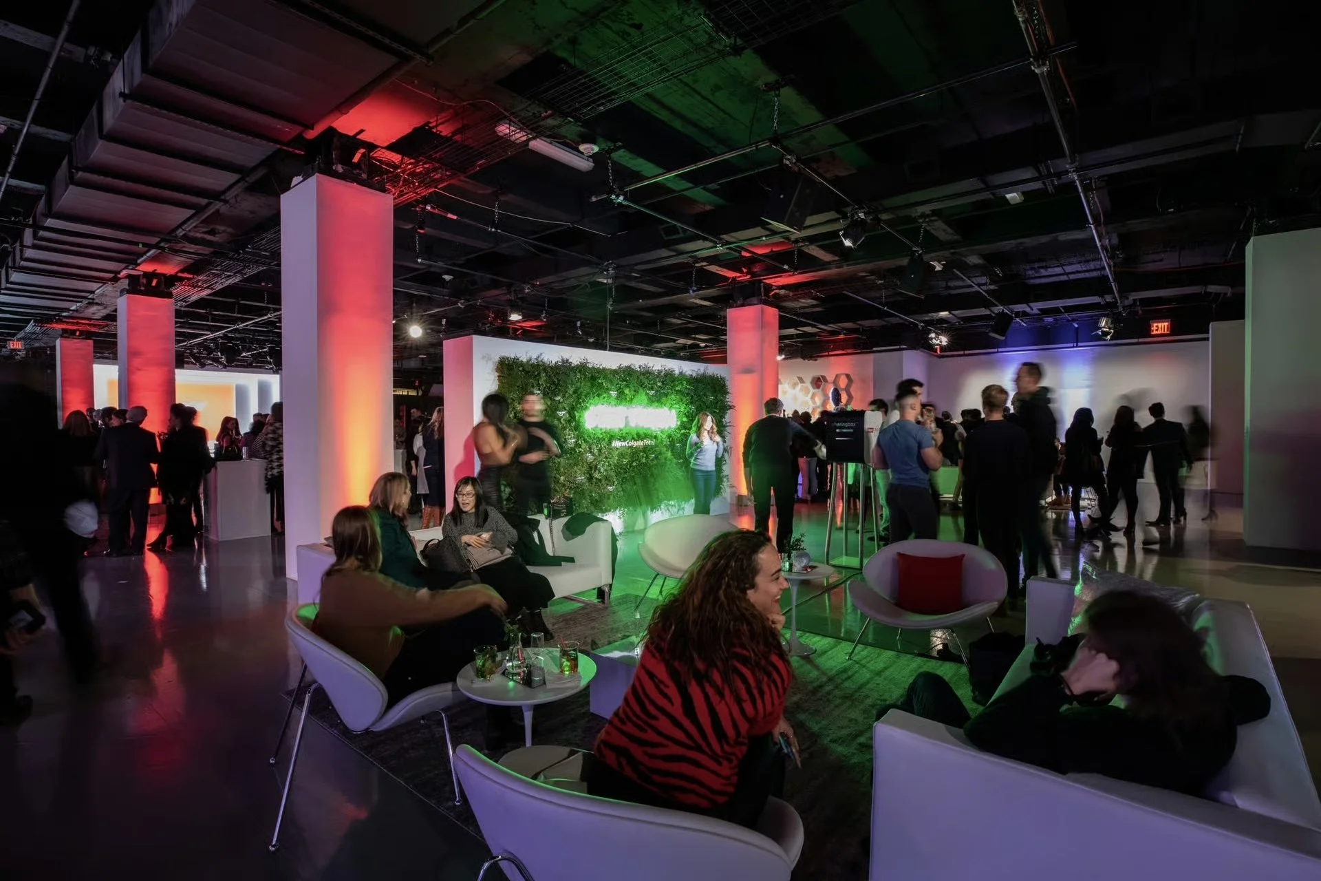 People socializing and sitting on white couches in a modern, dimly-lit lounge with colorful accent lighting, a green wall with illuminated sign, in an event space or gallery.