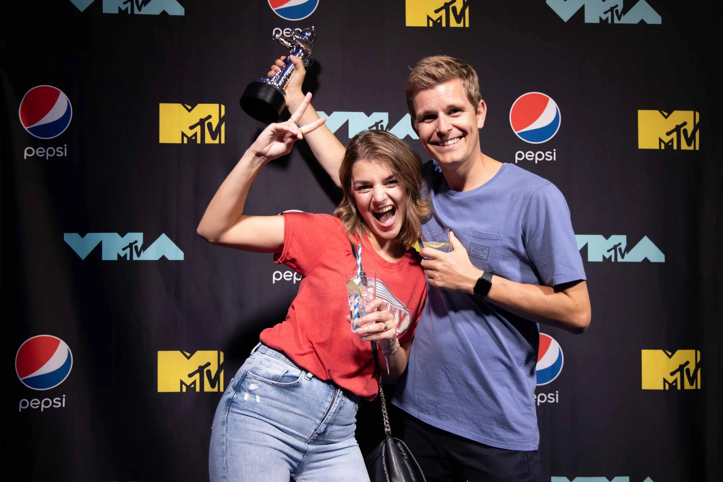 Two young adults celebrating at the MTV event, with one woman holding a trophy and both smiling; the woman is wearing a red t-shirt and jeans, the man is in a blue t-shirt; they are posing in front of a black backdrop with MTV and Pepsi logos.
