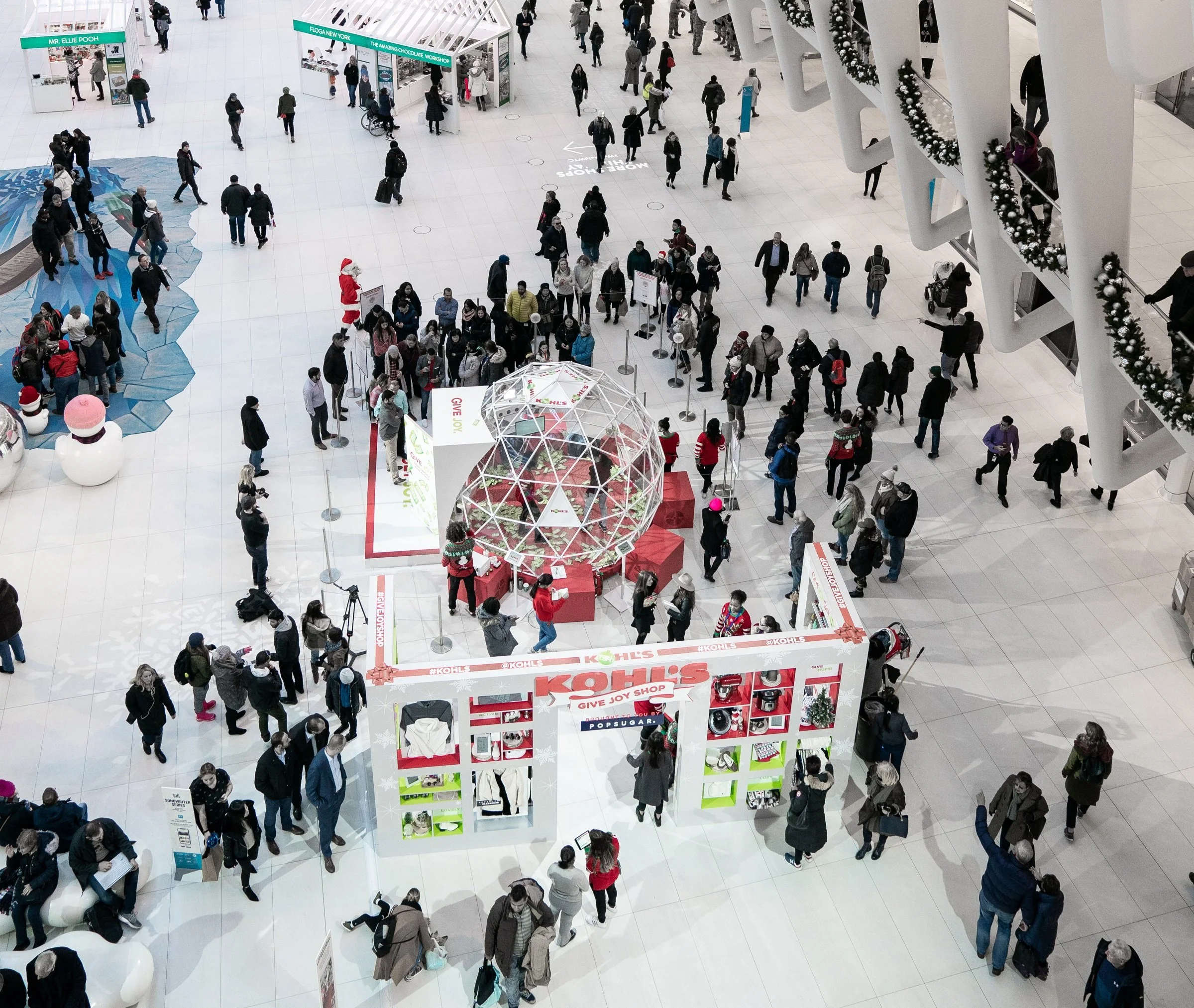 Crowd of people at a holiday-themed shopping mall event with a KOHL'S gift joy shop display, Christmas decorations, and a glass dome centerpiece.