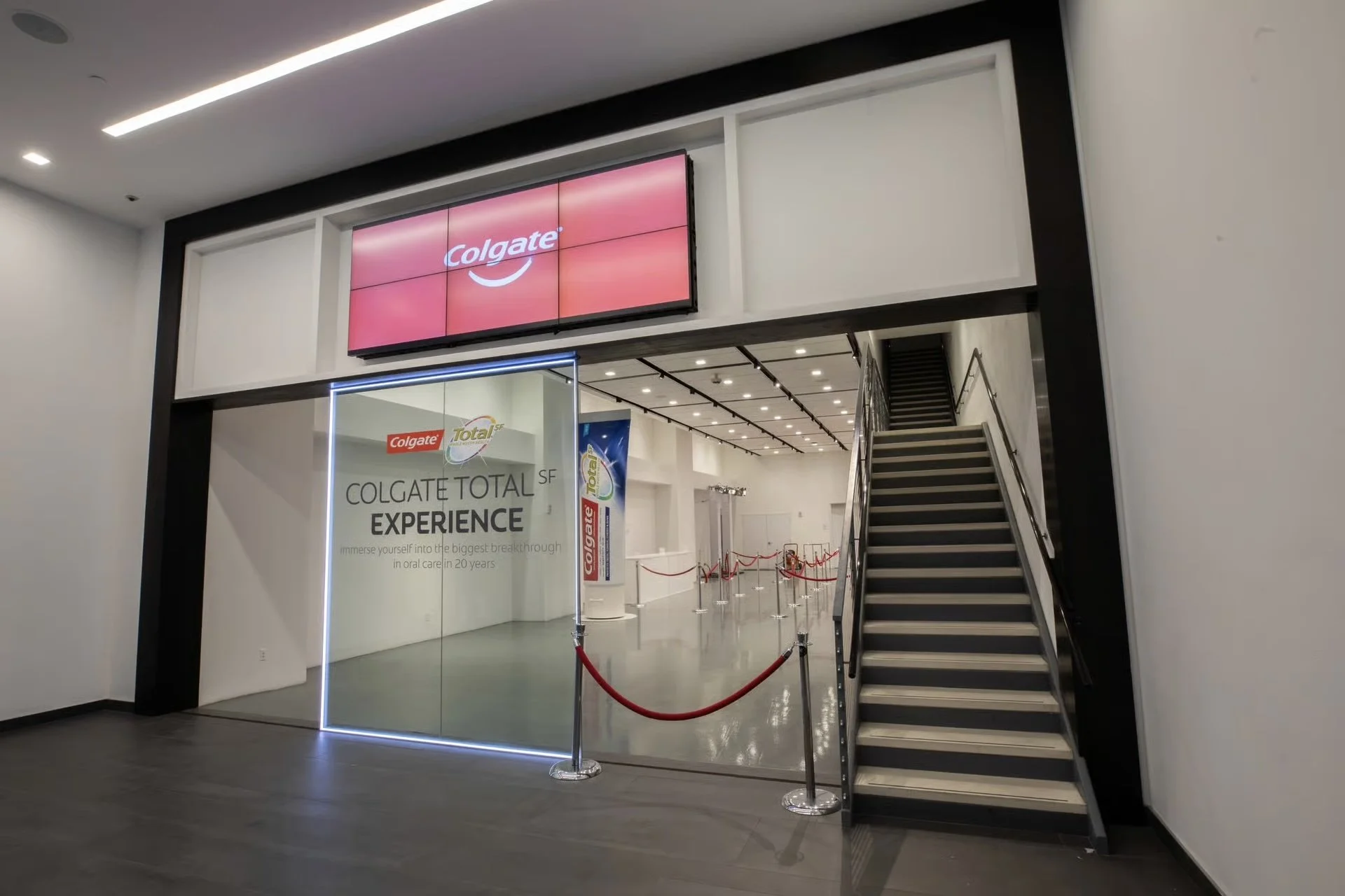 Entrance to the Colgate Total SF Experience exhibit with a glass door, red velvet ropes, and a staircase on the right side, inside a modern building.