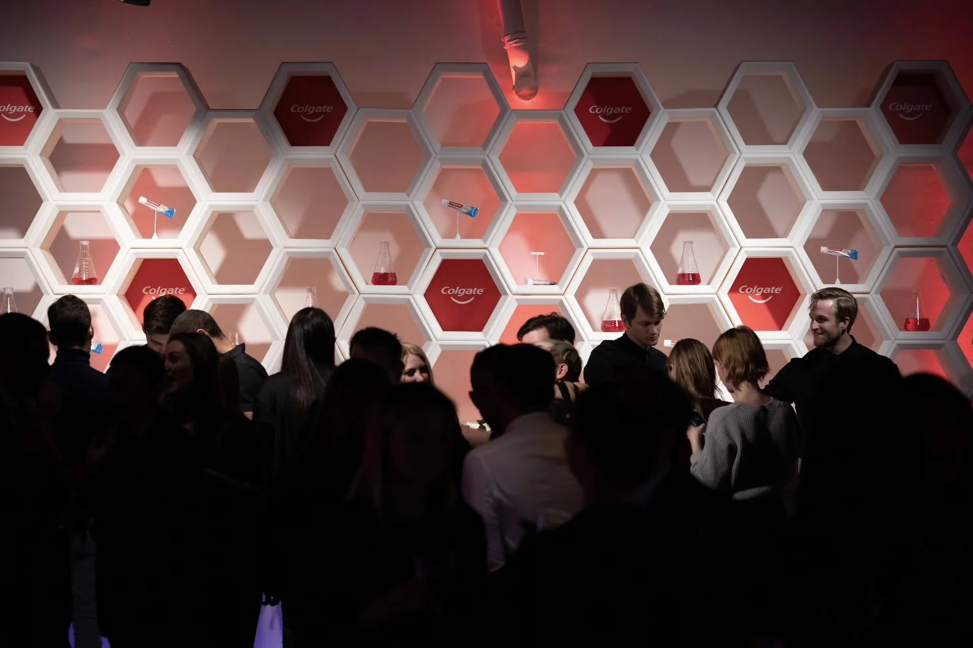 A crowd of people at an event with a decorative honeycomb wall featuring Colgate branding and laboratory glassware with red liquid.