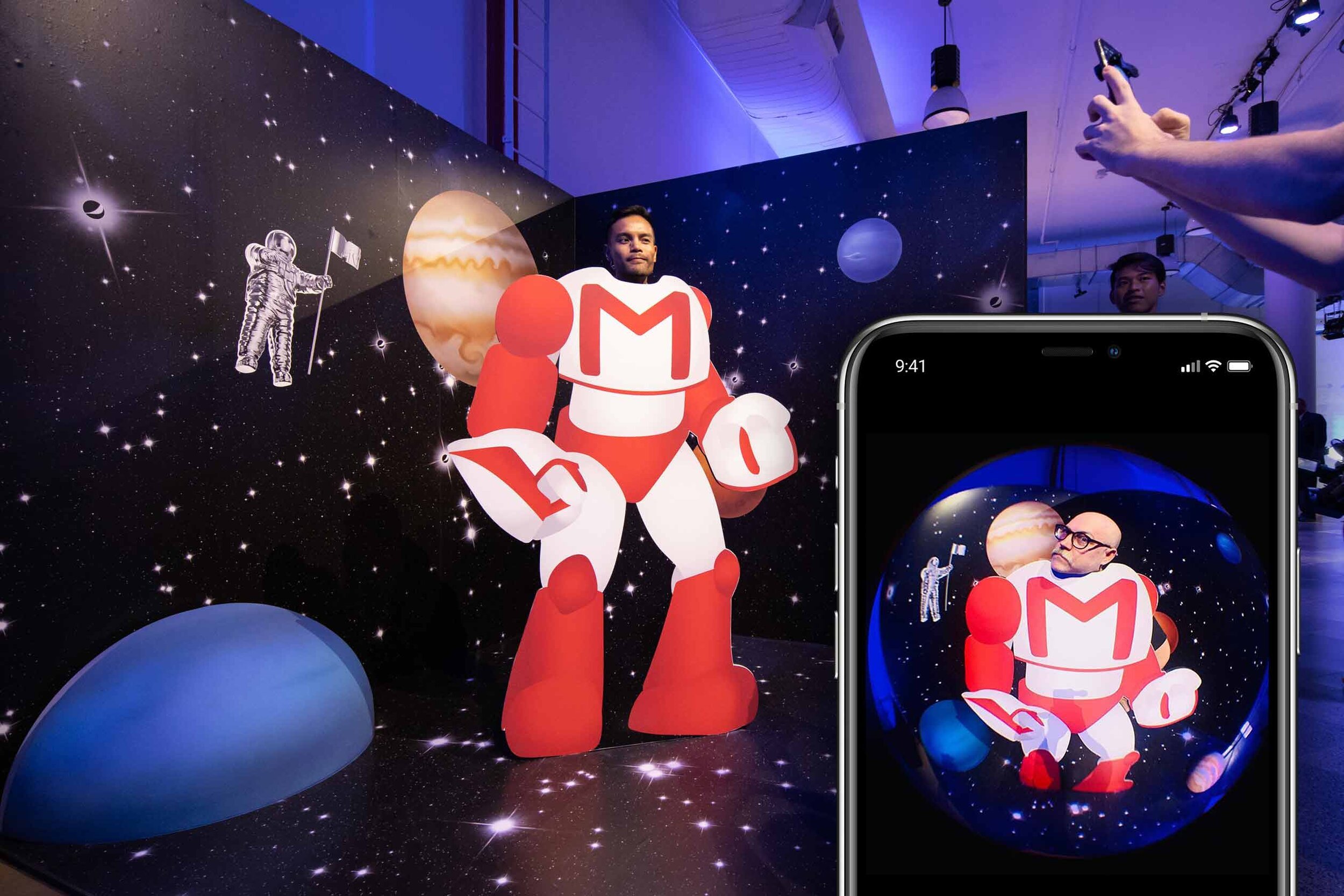 A person dressed as a robot with the Gmail logo on its chest is standing in front of a space-themed backdrop featuring planets and astronauts. Someone is taking a photo of them with a smartphone. The scene is displayed on a large screen and within a 
