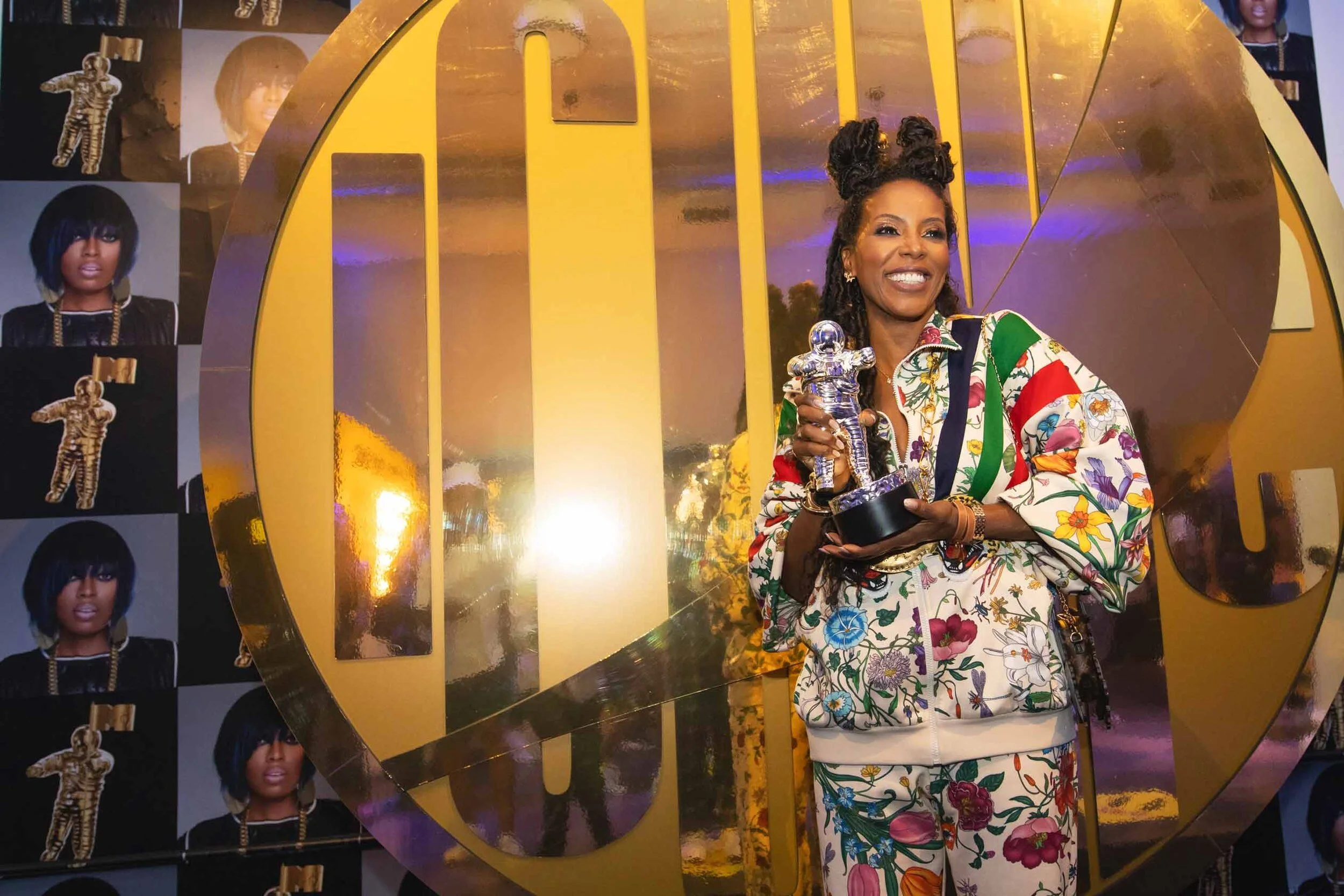 Woman with dark skin, wearing a colorful floral patterned tracksuit, holding a silver trophy, standing in front of a large golden logo at an event.