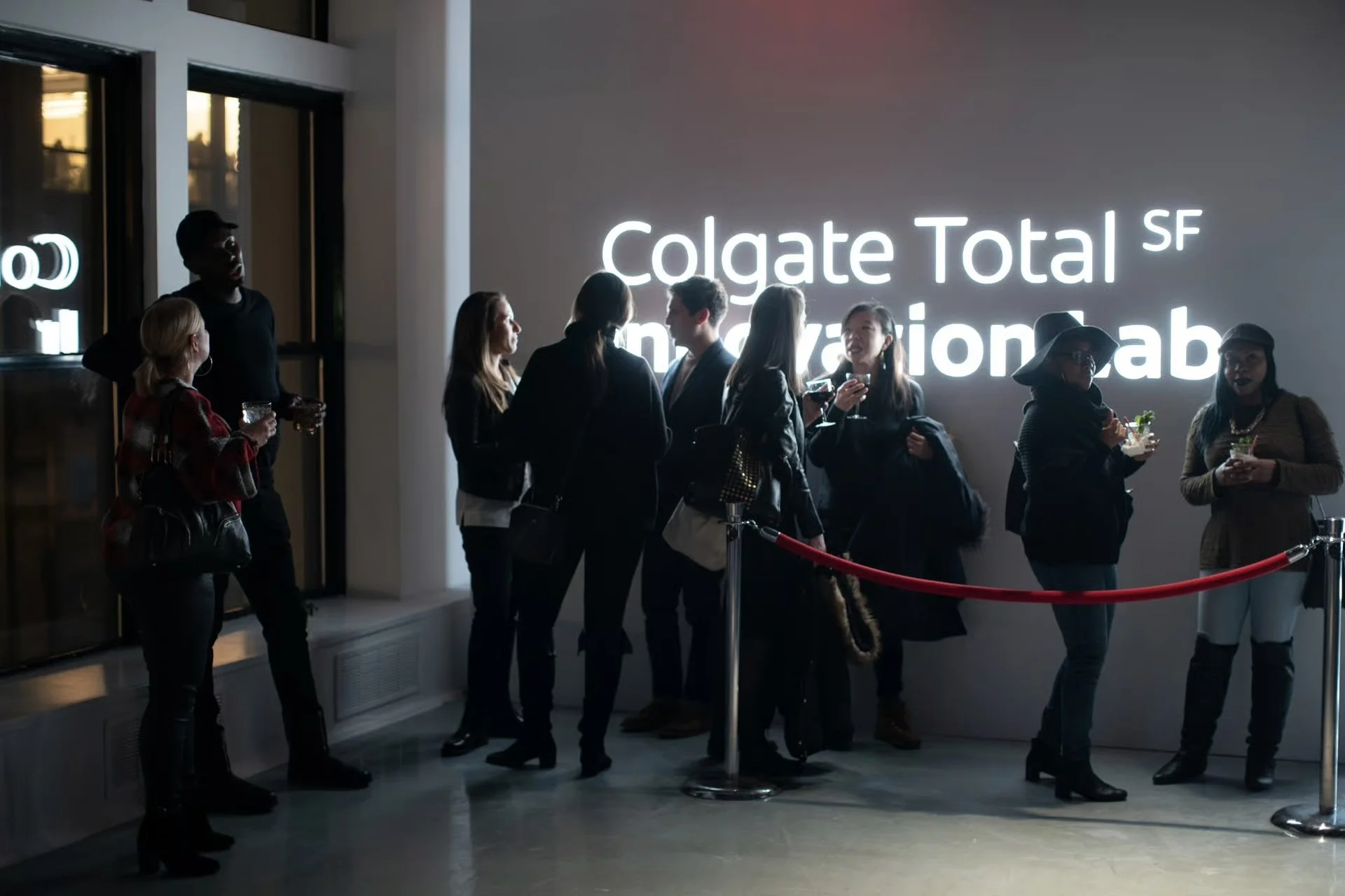 Group of people standing in line at an event, drinking and chatting in front of a large illuminated sign that reads 'Colgate Total SF' and other partially visible text.