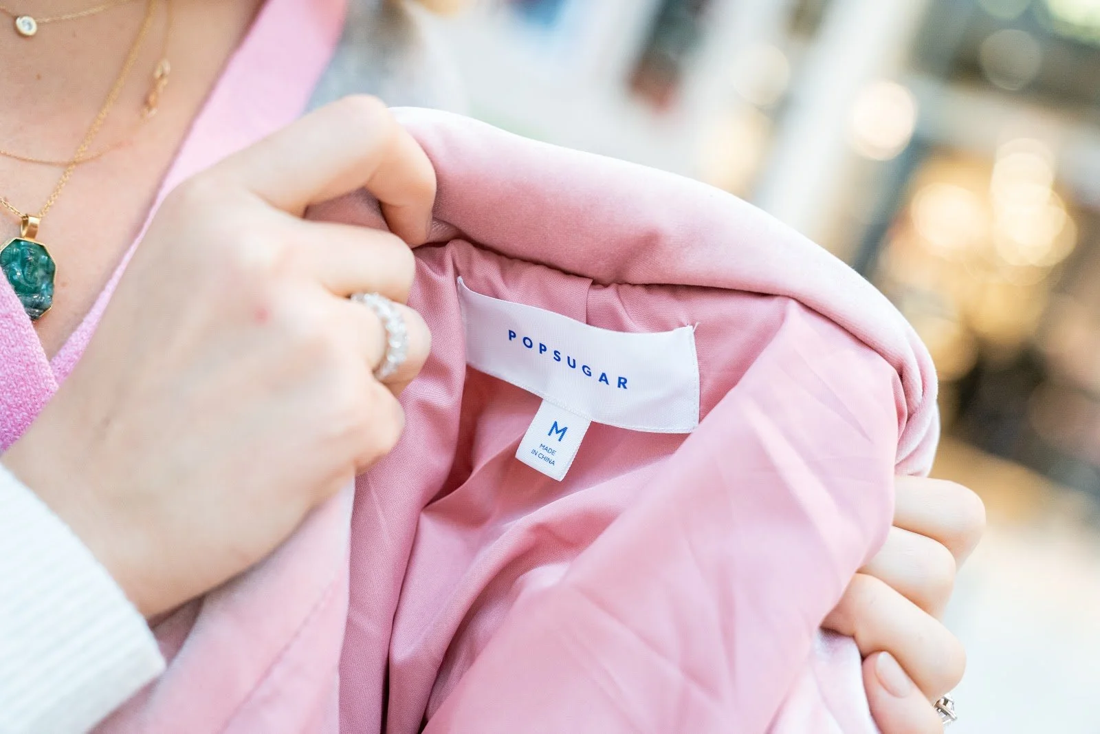 A person holding open a pink PopSUGAR jacket to show the label, which indicates it's a size M and made in China.