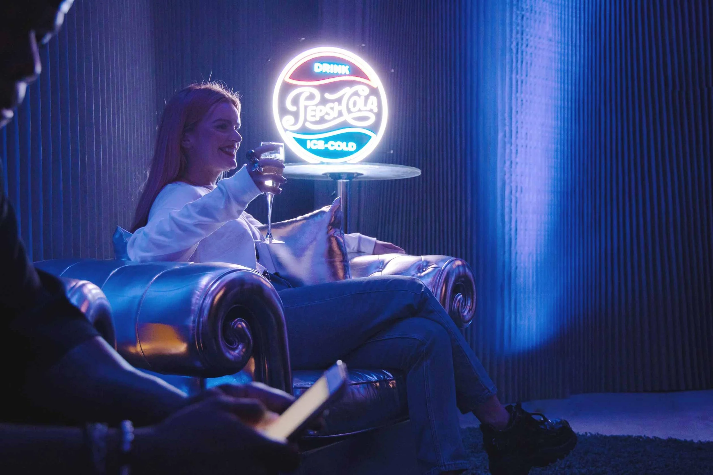 Woman sitting on a silver leather couch drinking a glass of champagne, with a neon Pepsi-Cola sign behind her in a dark, blue-lit room.