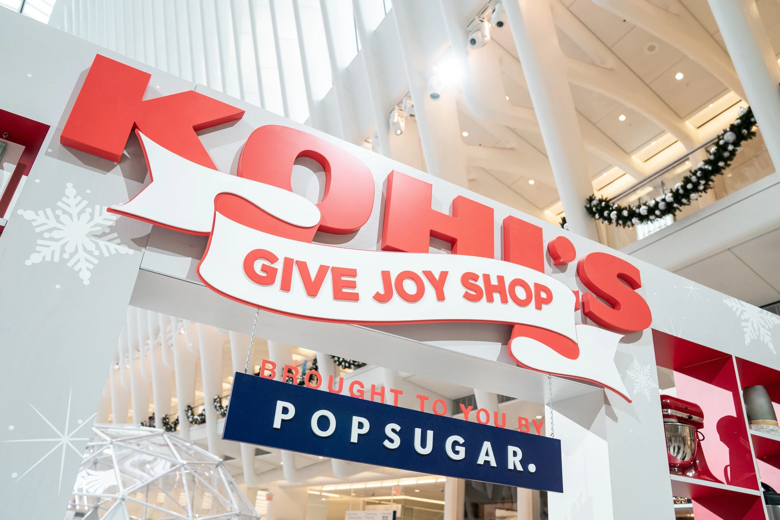 Holiday decorated gift shop sign with red and white lettering that reads 'Give Joy Shop' and 'Brought to you by Popsugar' inside a shopping mall.