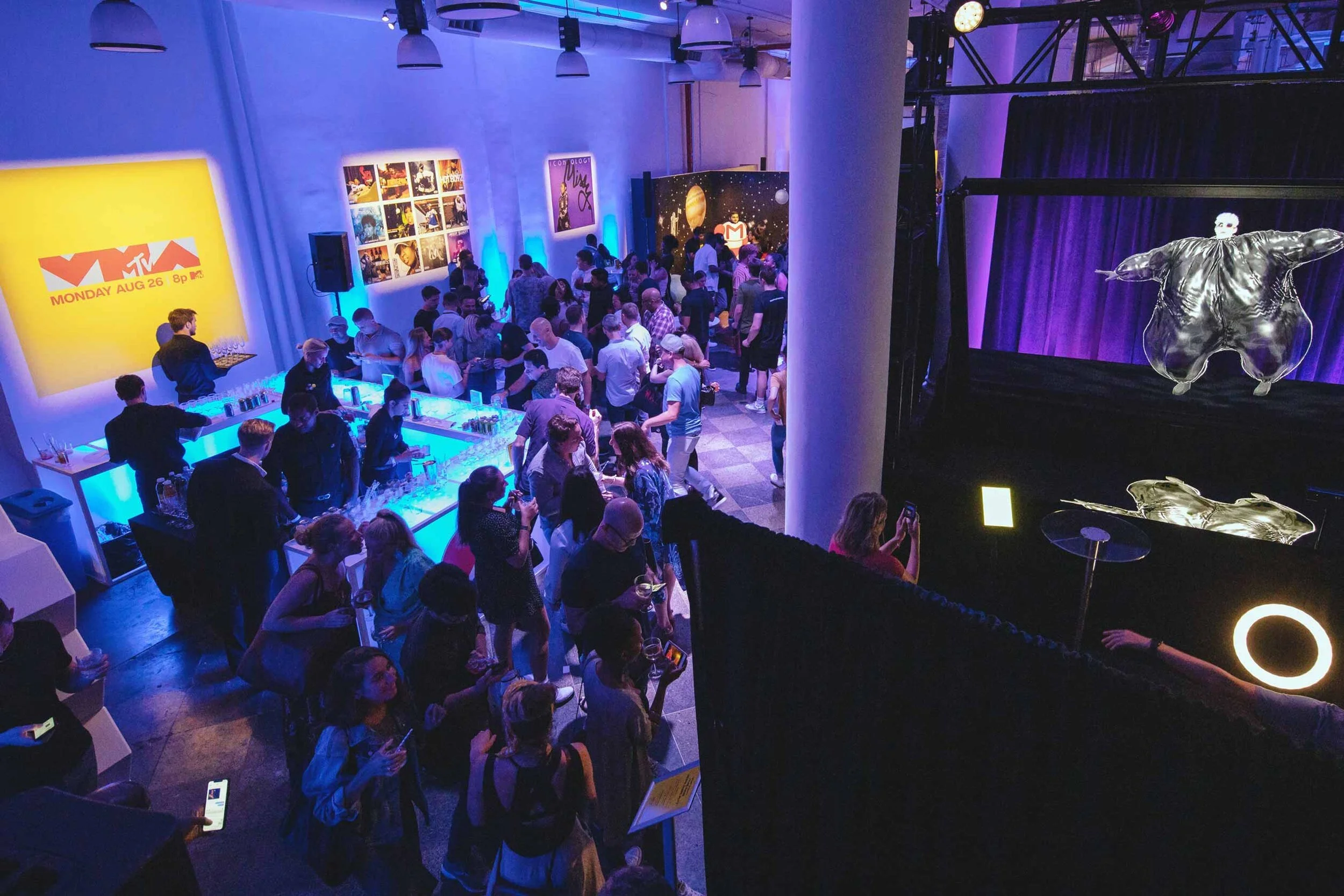 A crowded indoor event with a blue and purple lighting theme, a bar area with bartenders serving drinks, and a large screen displaying the MTV logo and a date. Some people are standing in line at the bar, others are socializing and taking photos, wit