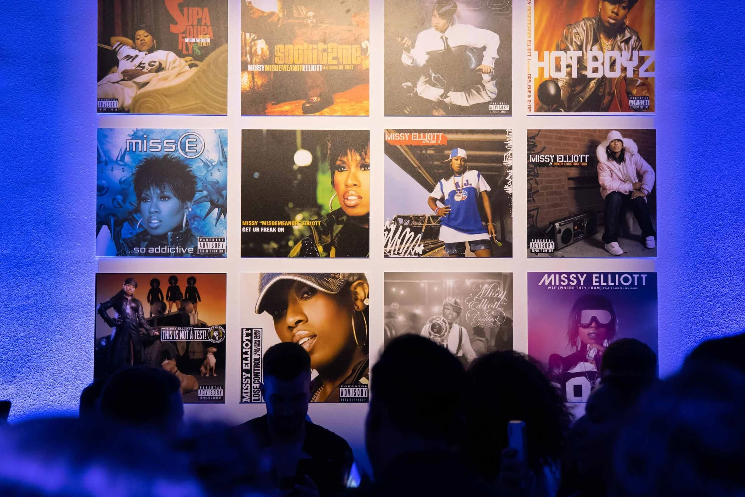 A wall displays a grid of twelve album covers by Missy Elliott, featuring her images, artwork, and album titles, with people gathered in front of the display.