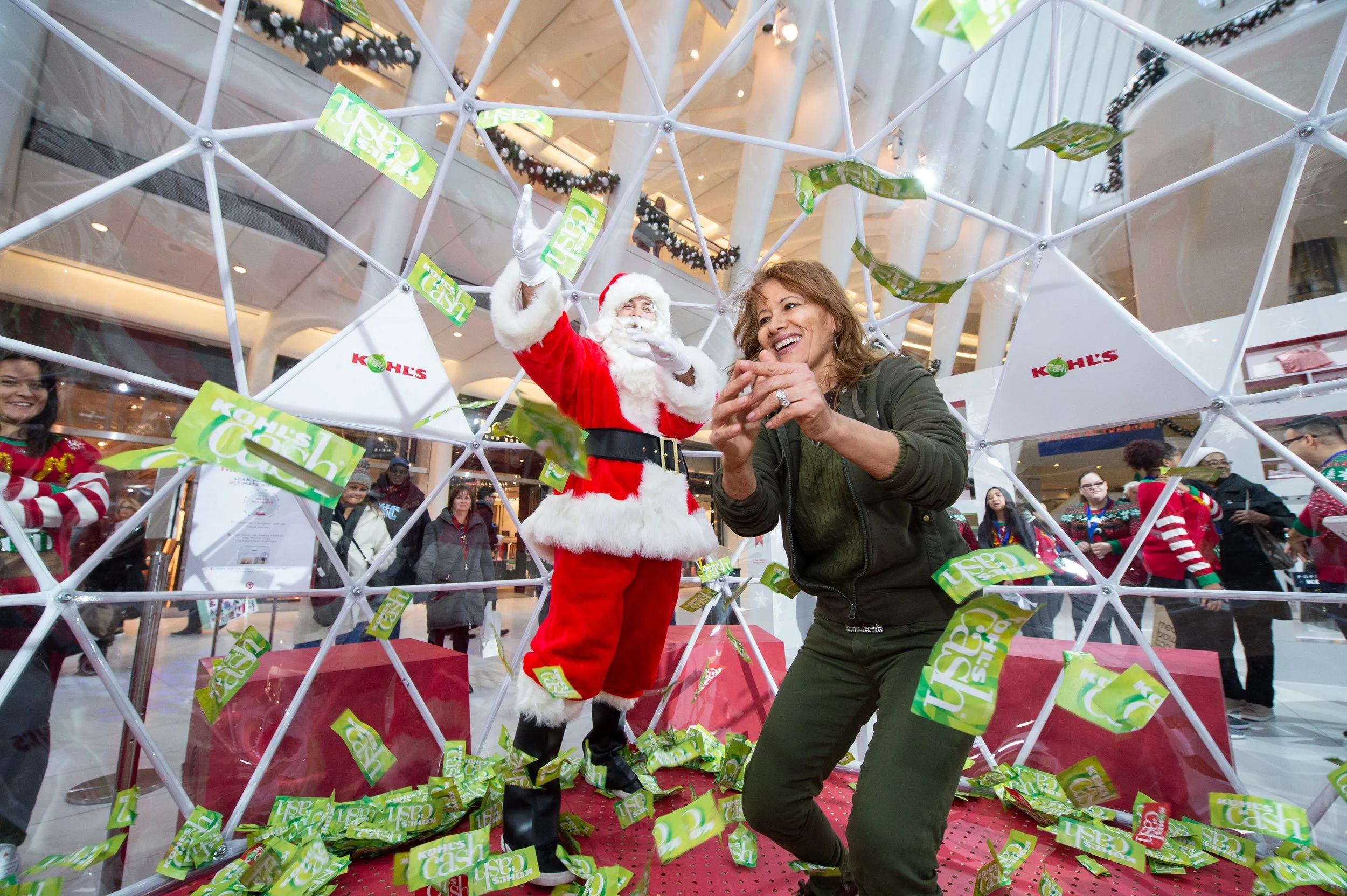 People celebrating Christmas in a shopping mall with a Santa Claus inside a gift box frame, surrounded by falling green Kohls cash notes, with smiling onlookers. One woman is leaning into the frame, smiling joyfully.