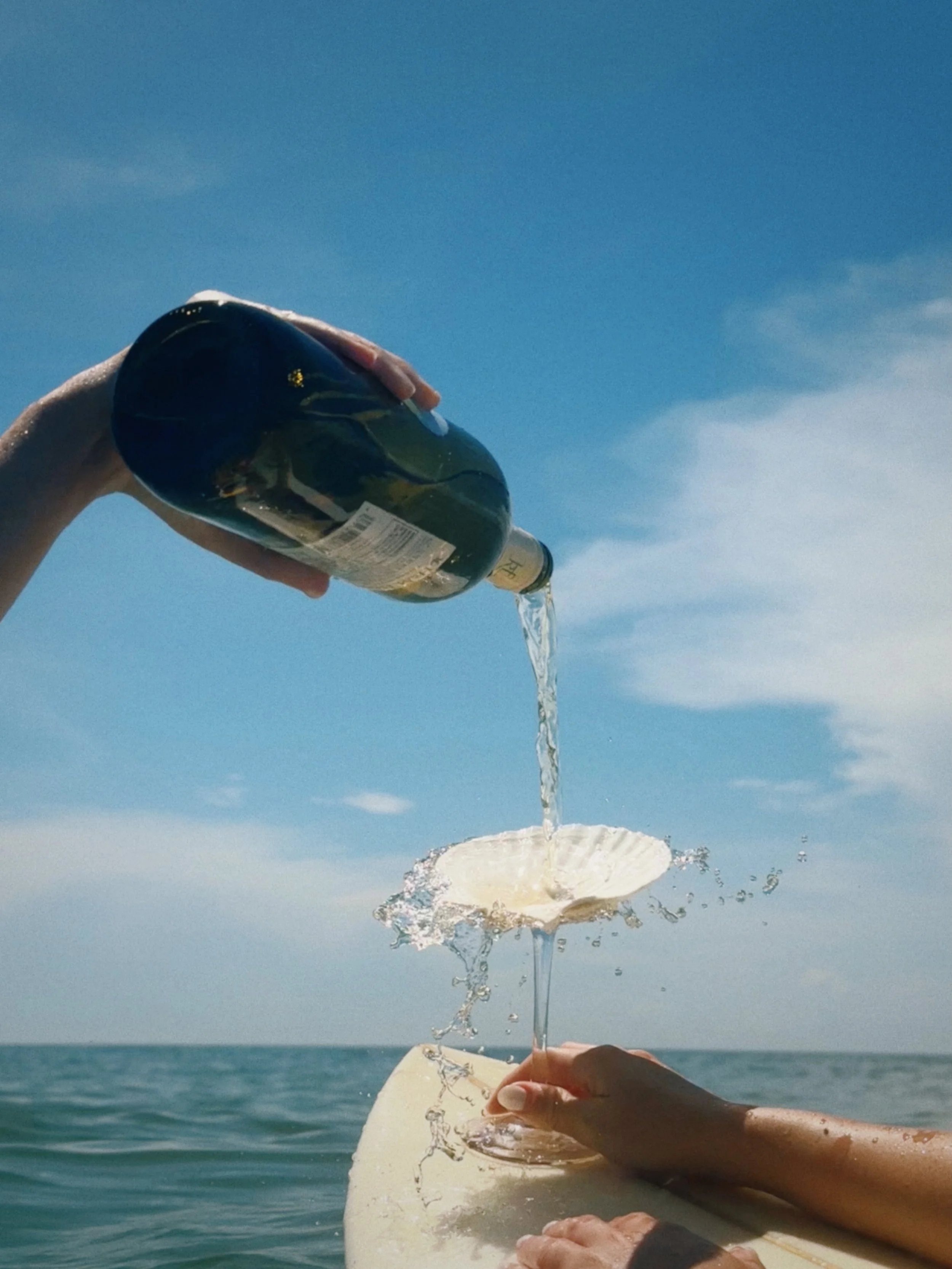 Two hands pouring sparkling wine into a shell-shaped glass on a surfboard, with ocean and blue sky in the background.