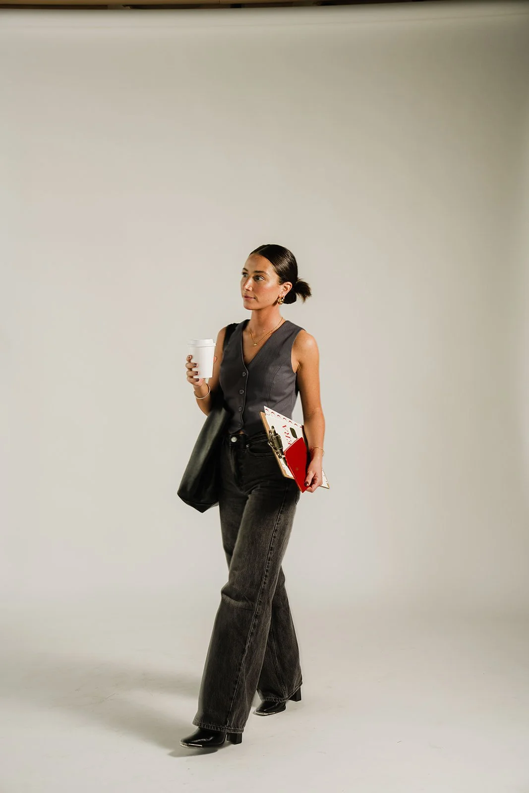 A woman dressed in a sleeveless gray vest, black jeans, and black boots, holding a coffee cup and some notebooks or folders, walking against a plain white background.