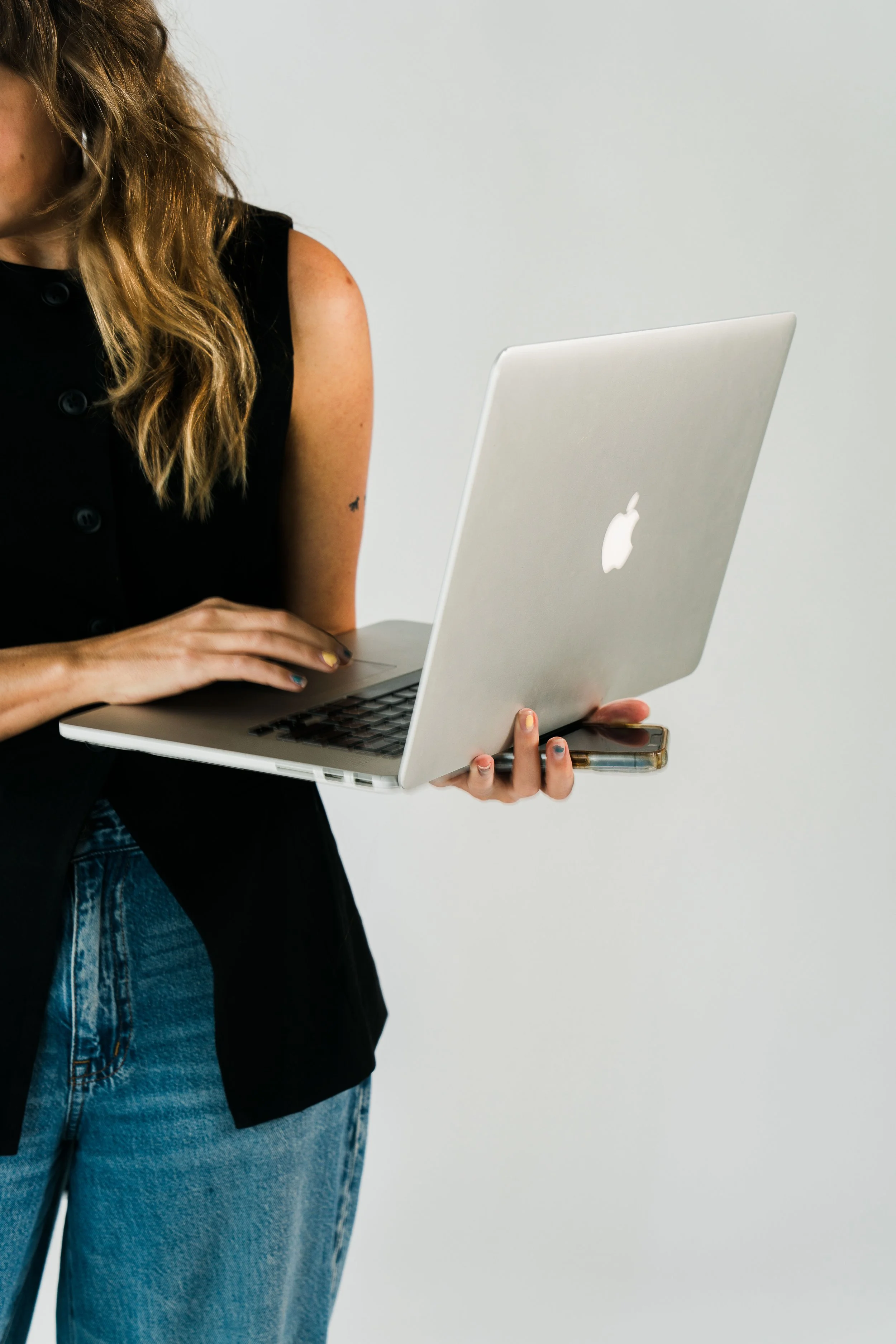 A woman with wavy blond hair wearing a black vest and blue jeans, holding a silver Apple MacBook laptop in her left hand and a smartphone in her right hand, against a plain white background.