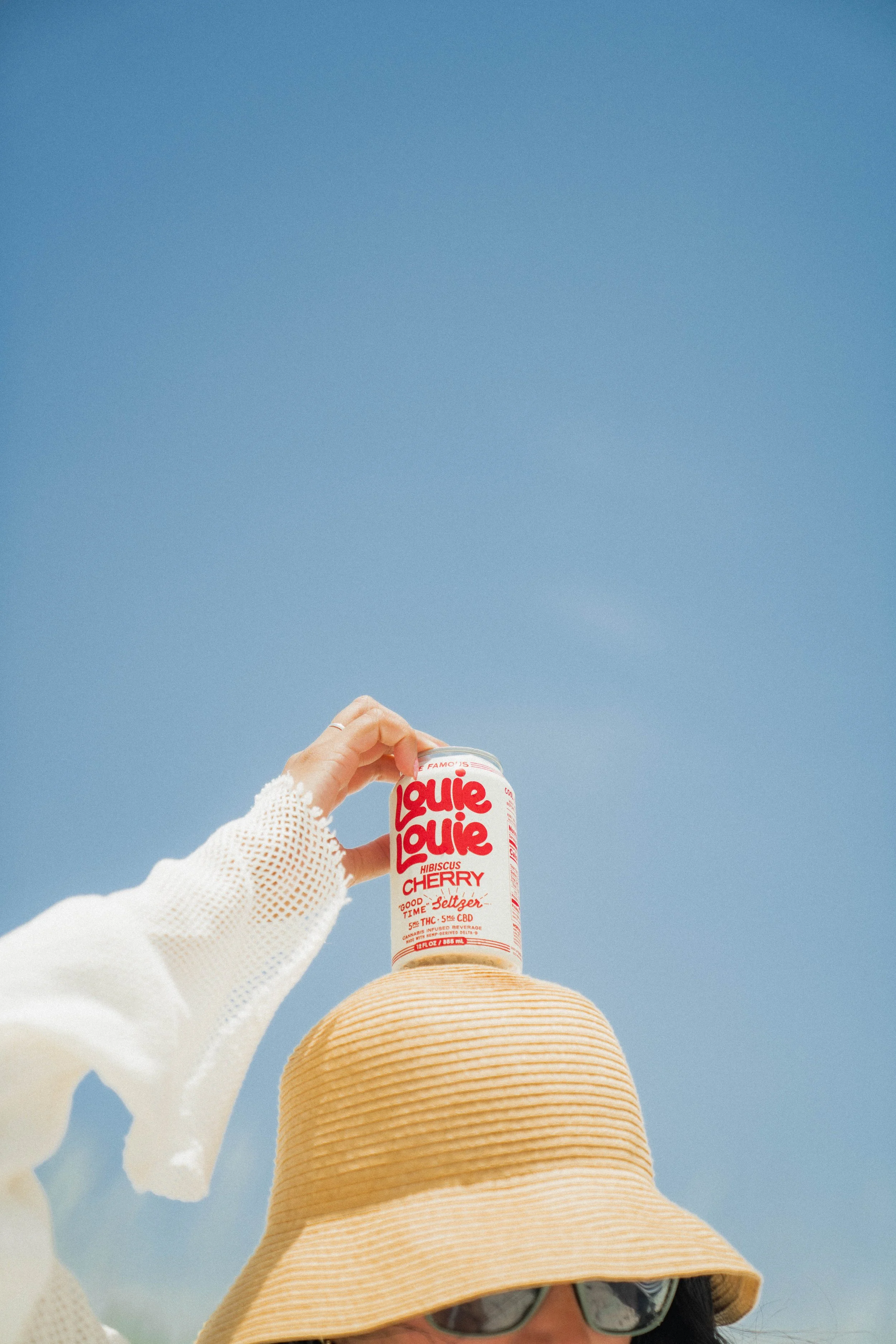 Person wearing a large, wide-brimmed straw hat and sunglasses, holding a can of Louie Louie cherry flavored beverage against a clear blue sky.