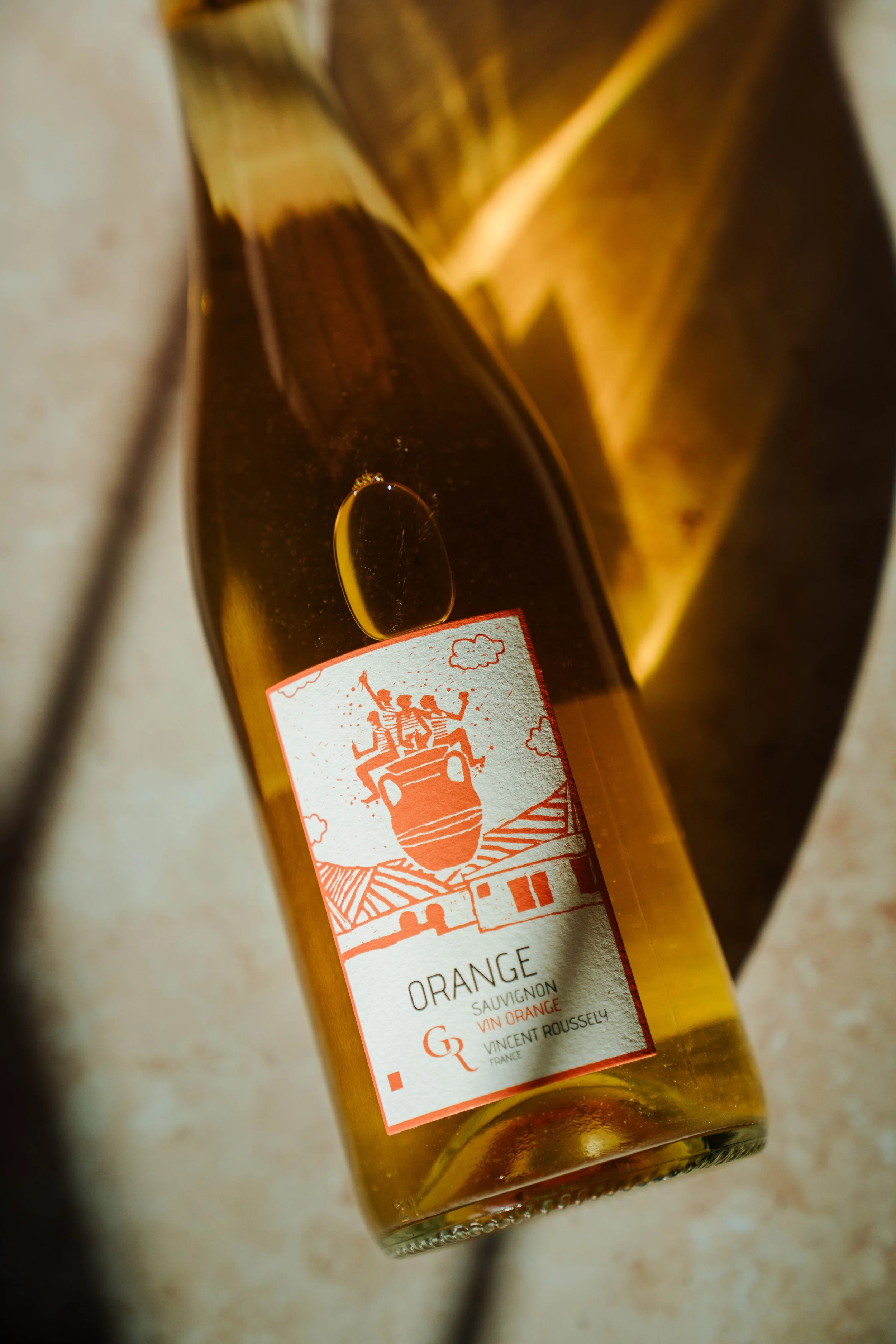 A bottle of orange wine with a label featuring an illustration of a vineyard landscape and three people carrying a large wine barrel, with the words 'Orange Sauvignon Vin Orange' and 'Vincent Roussely, France'.