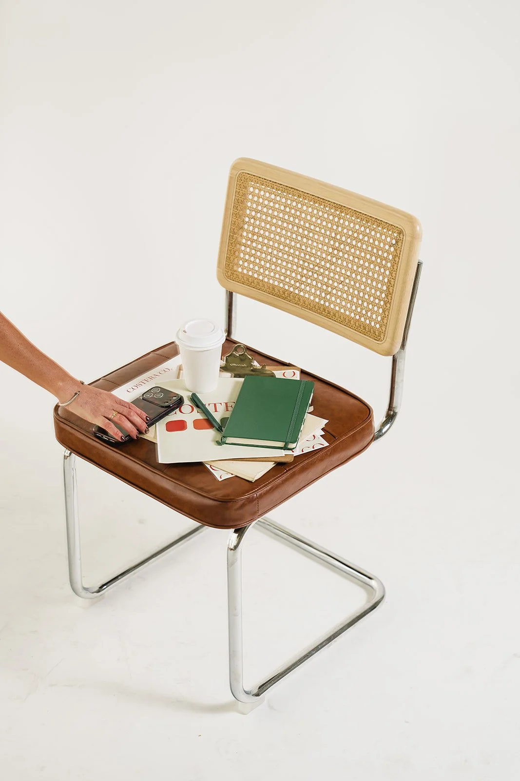 A brown leather chair with a woven rattan backrest, topped with a collection of papers, a green notebook, a pen, a black smartphone in a hand, a marker, and a disposable coffee cup with a white lid.