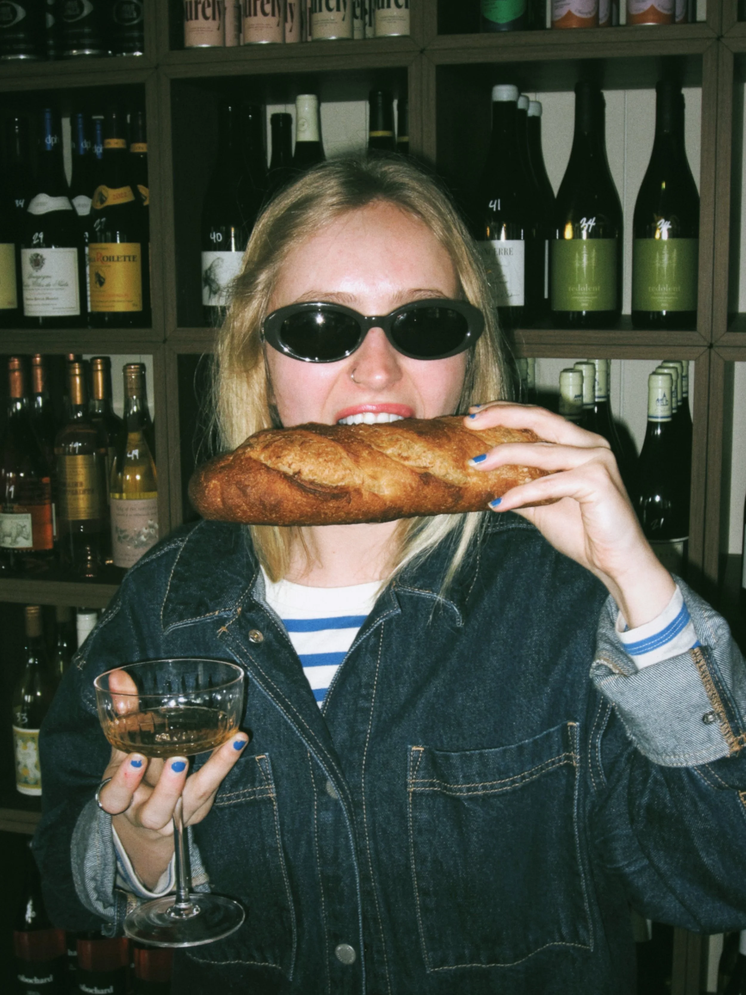 Woman with blonde hair wearing sunglasses and denim jacket holding a baguette in her mouth and a glass of wine in her hand in front of wine shelves.