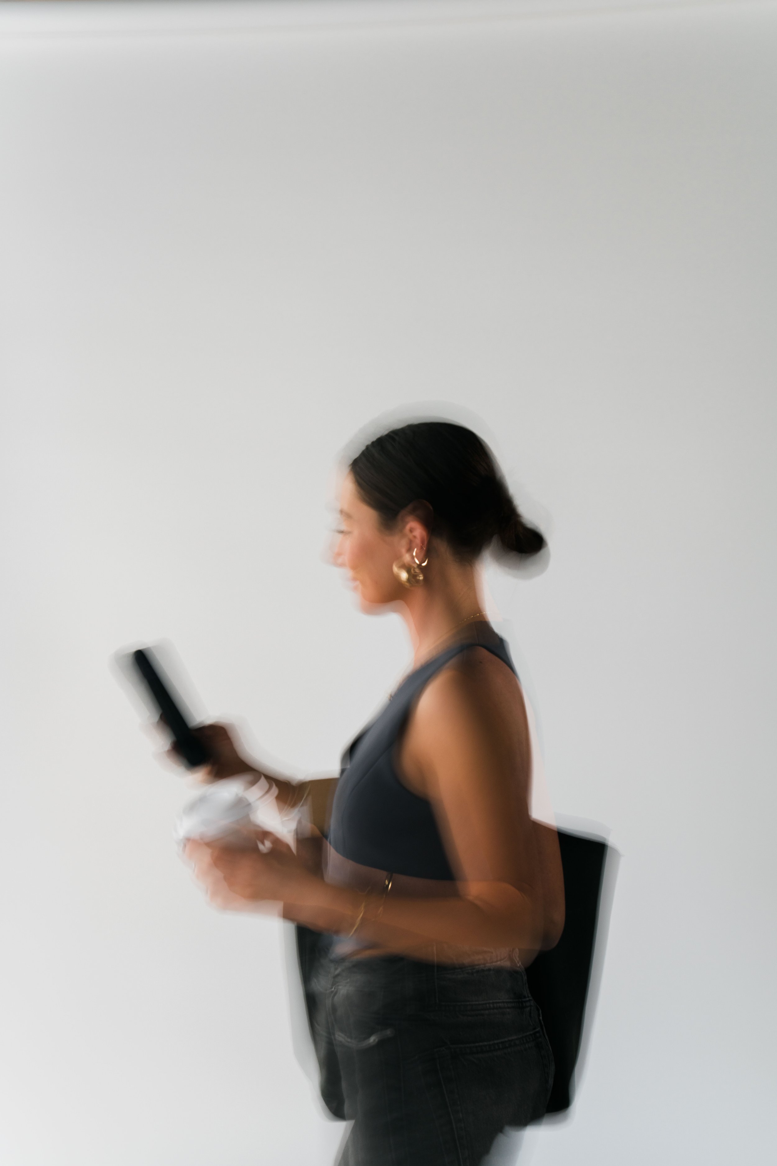 A woman with dark hair tied in a bun, wearing gold earrings and a black sleeveless top, is walking while looking at her phone, with a backpack on her back, against a plain white background.