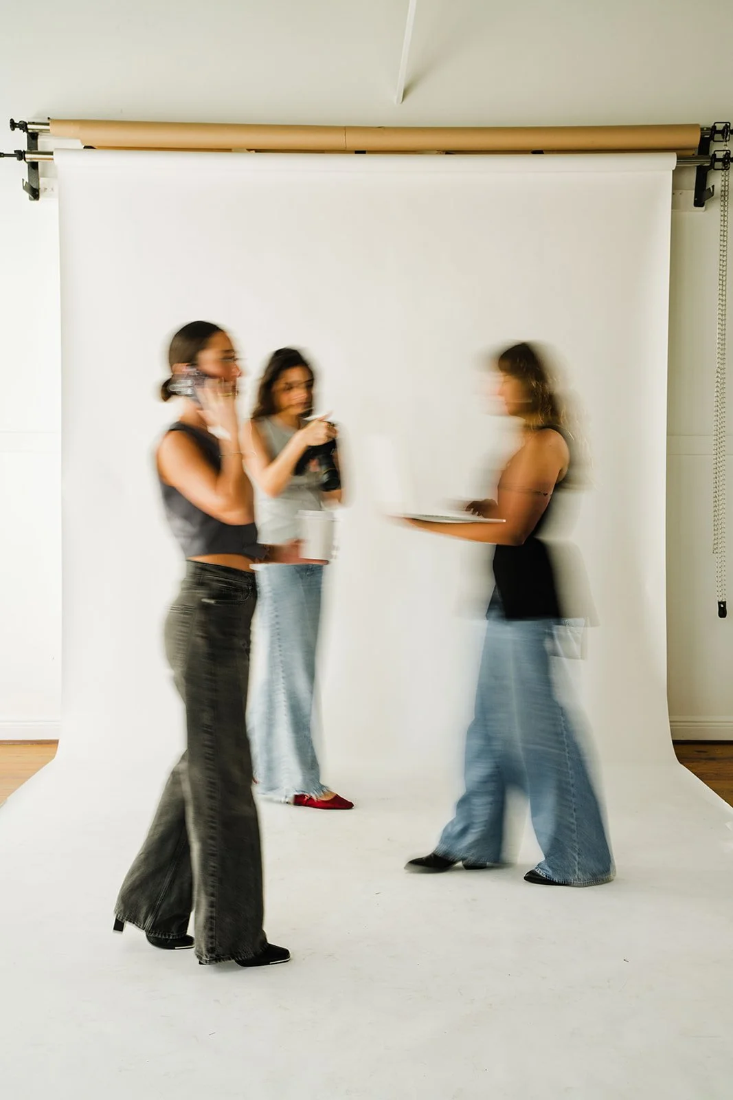 Three women in a photography studio with a white backdrop, one holding a tray, and the other two looking at her, with motion blur showing movement.